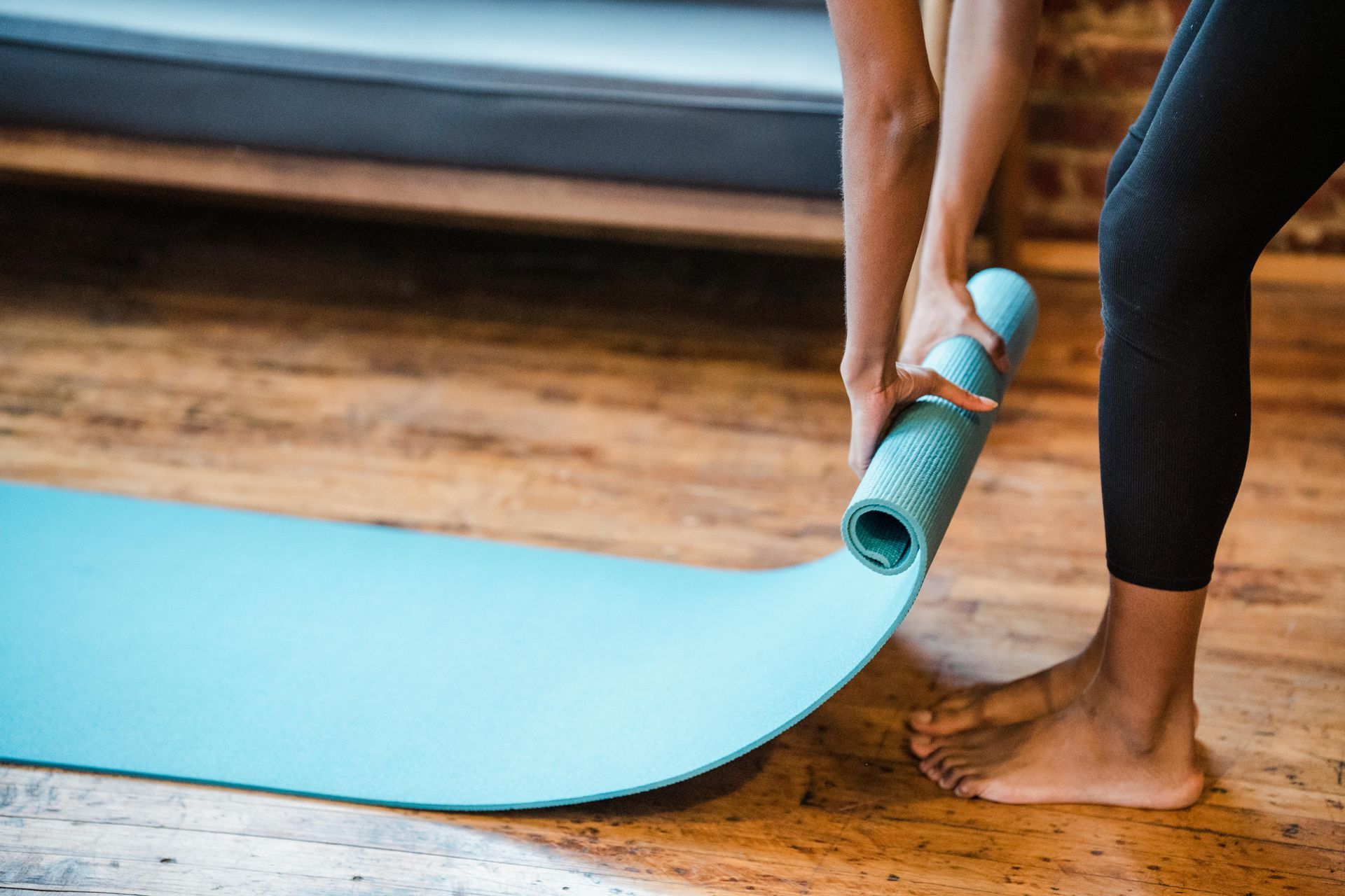 Person rolling out a blue yoga mat on a wooden floor in front of a brick wall.