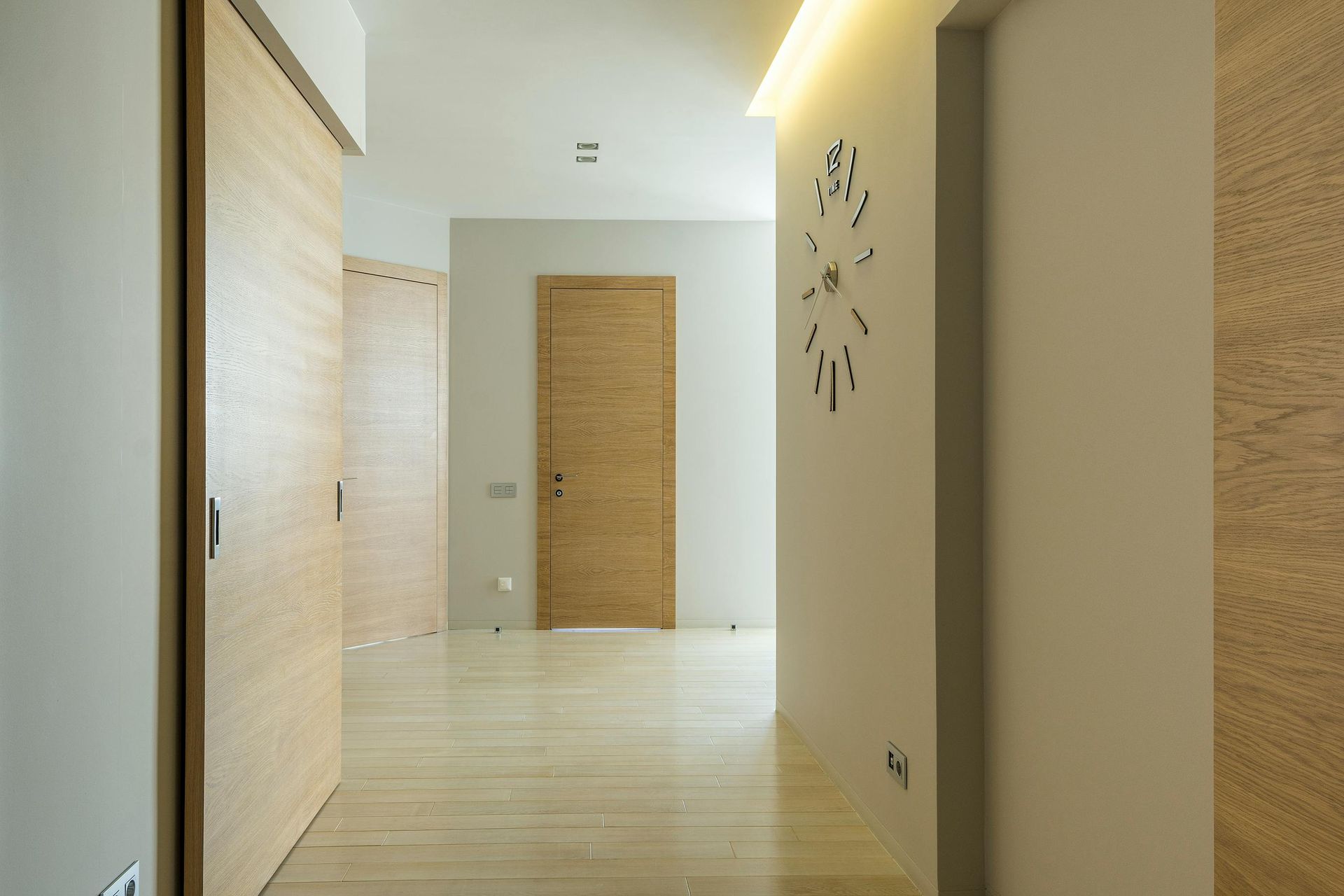 Long hallway with light wood doors and floors, neutral walls, and a wall clock.