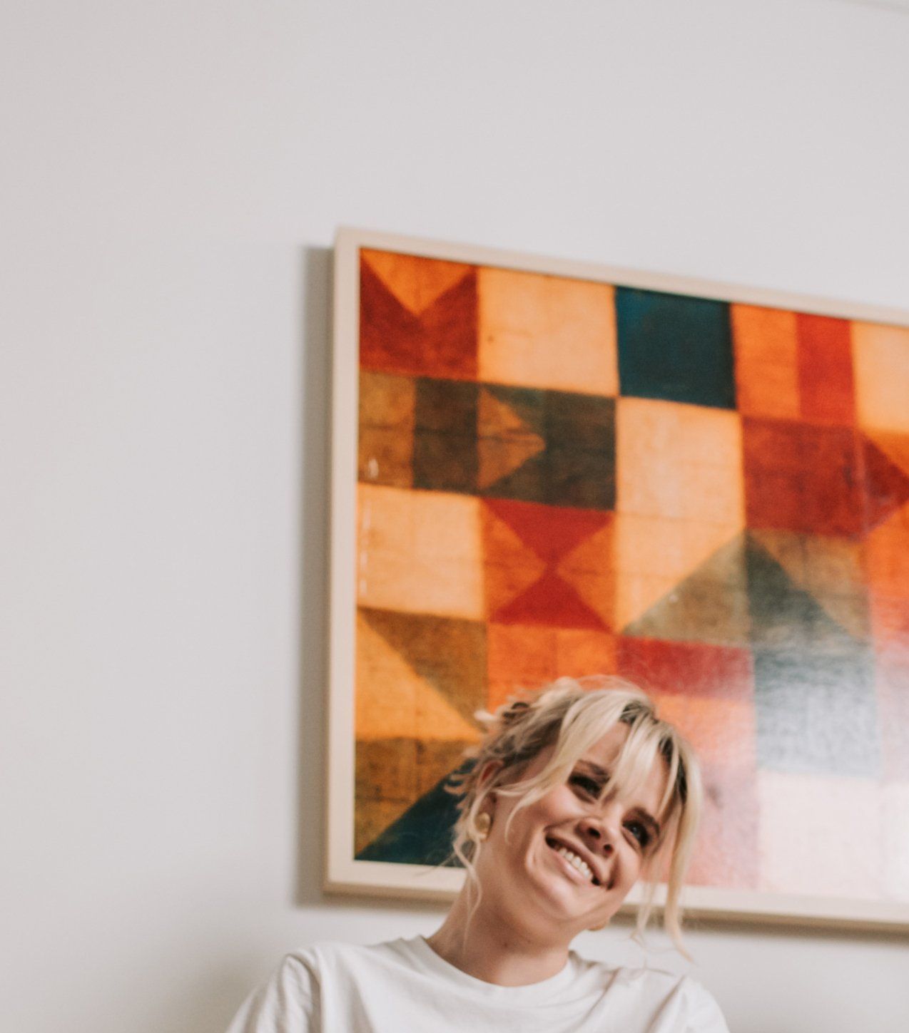 Smiling person with blonde hair in front of abstract art on a white wall.
