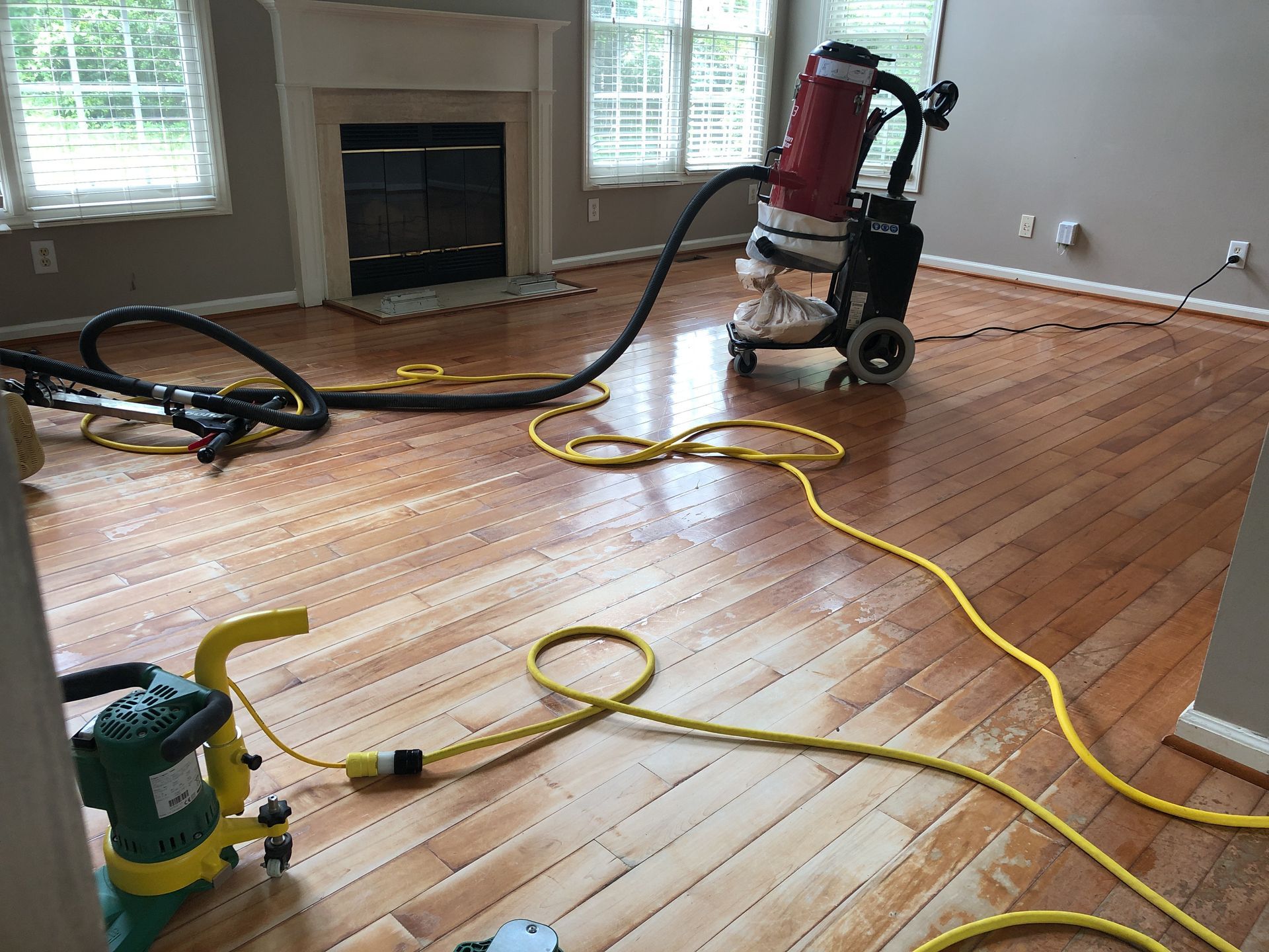 Floor sanding in progress: machine on wood floor, connected to vacuum. Fireplace and windows in background.