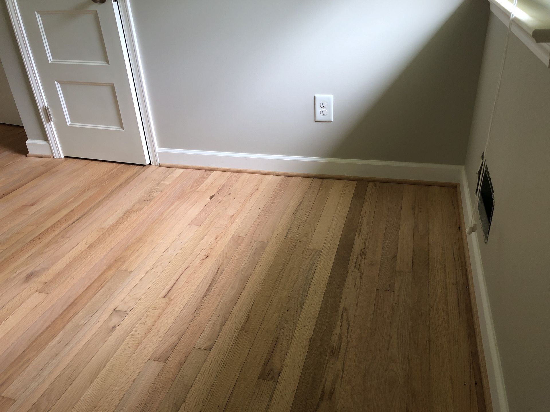 Hardwood floor in a room with light gray walls, a white door, and a wall outlet.