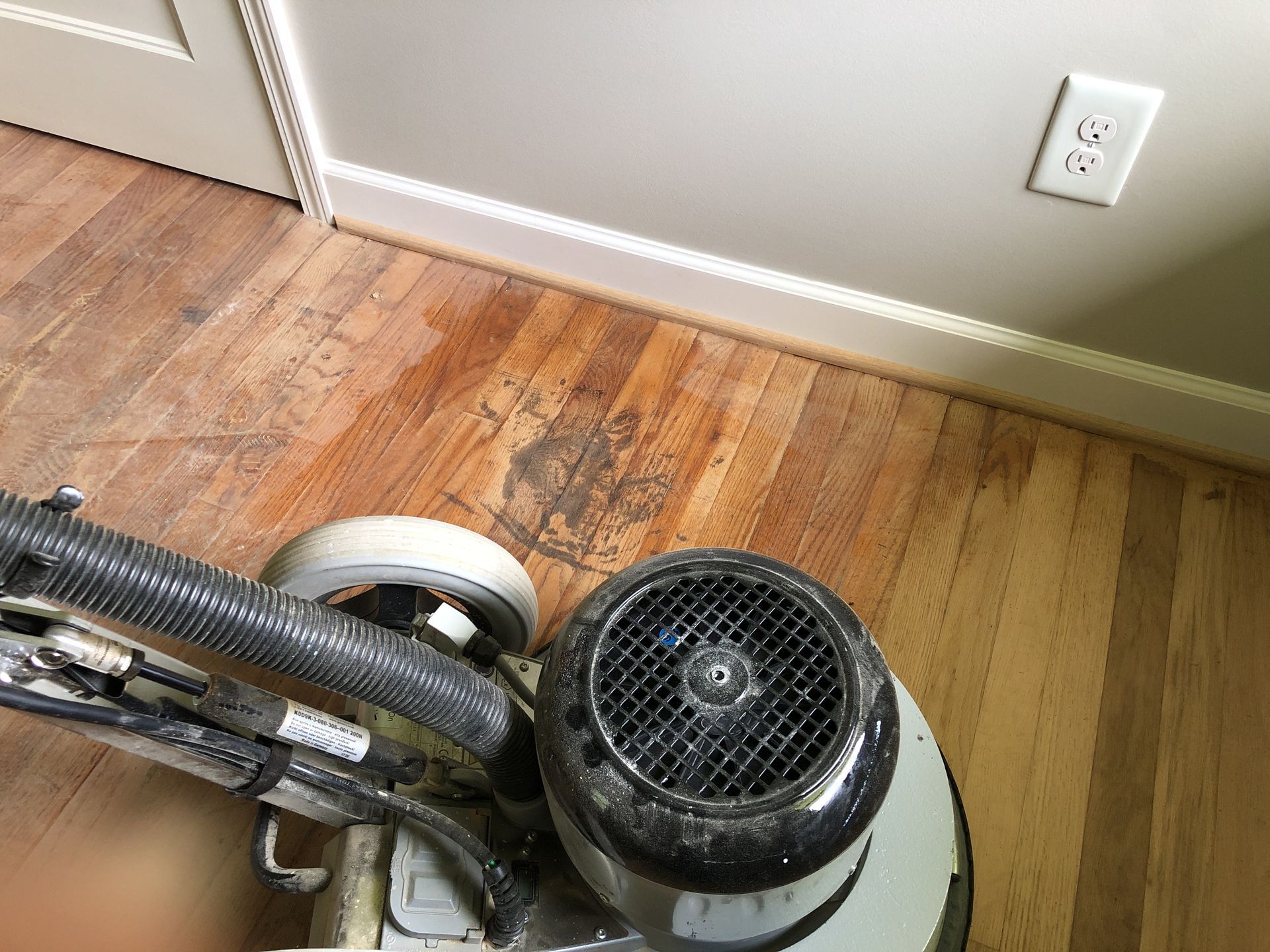 A floor sander in use on a wood floor next to a wall with baseboard and an electrical outlet.