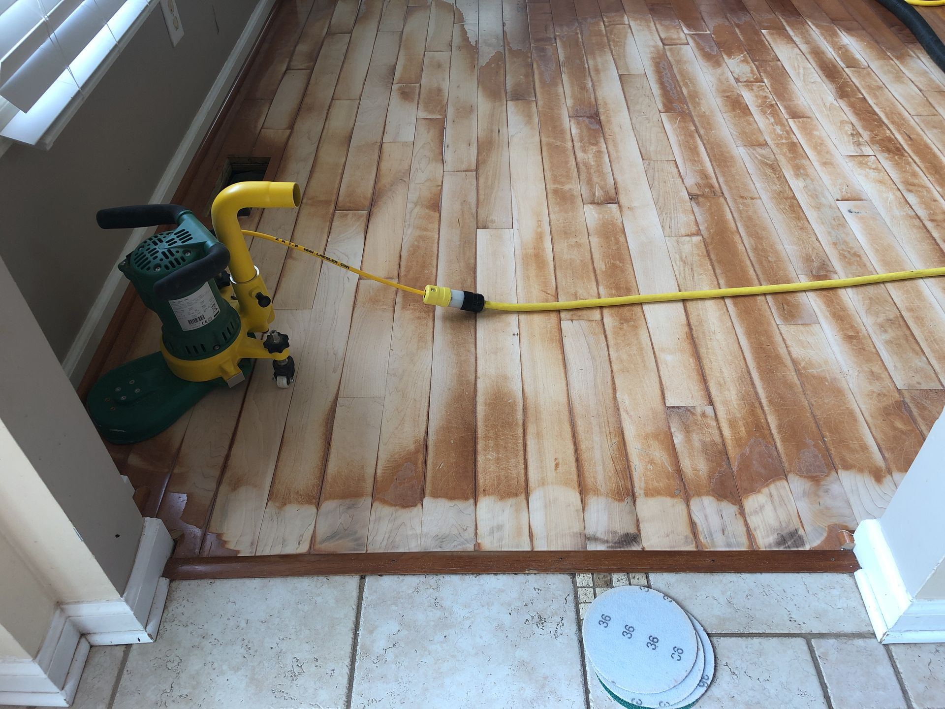 Floor being refinished with a yellow sanding machine. Wood floor has areas of bare wood, wet appearance.