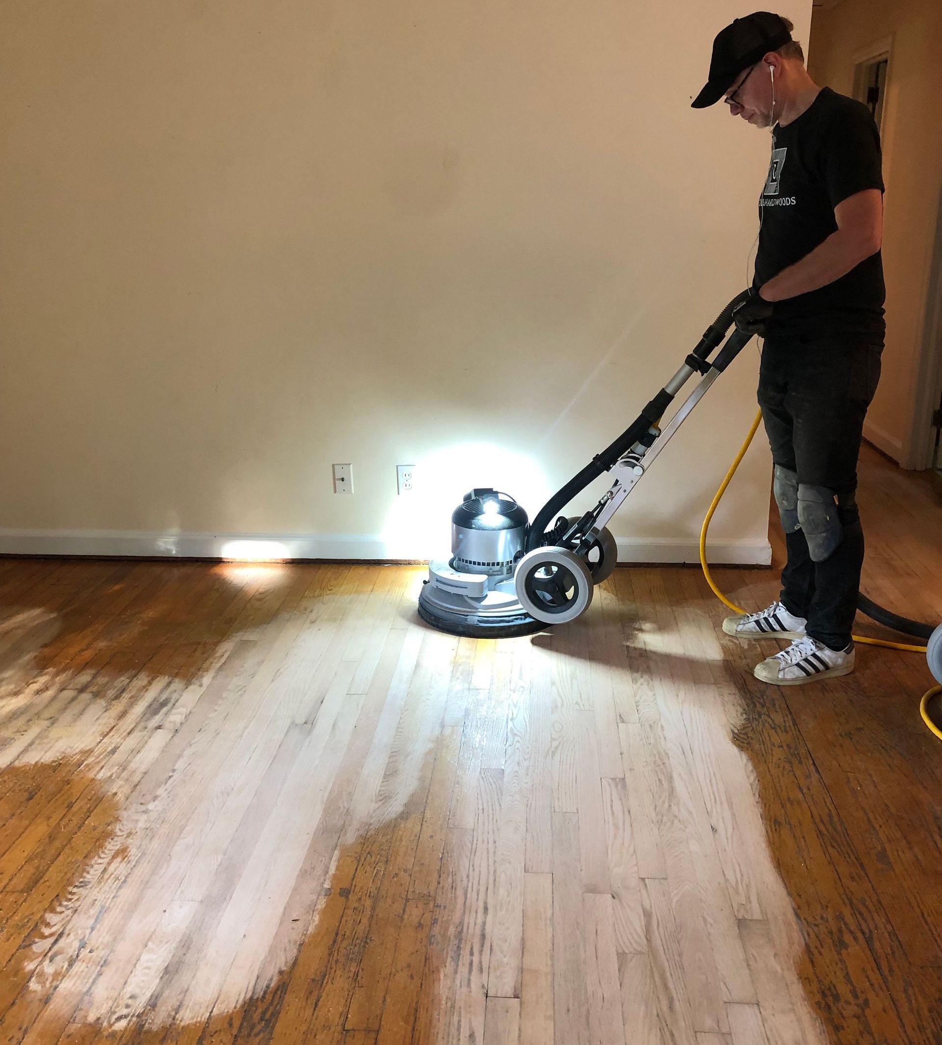 Person sanding a wooden floor with a floor sander in a room with light-colored walls.