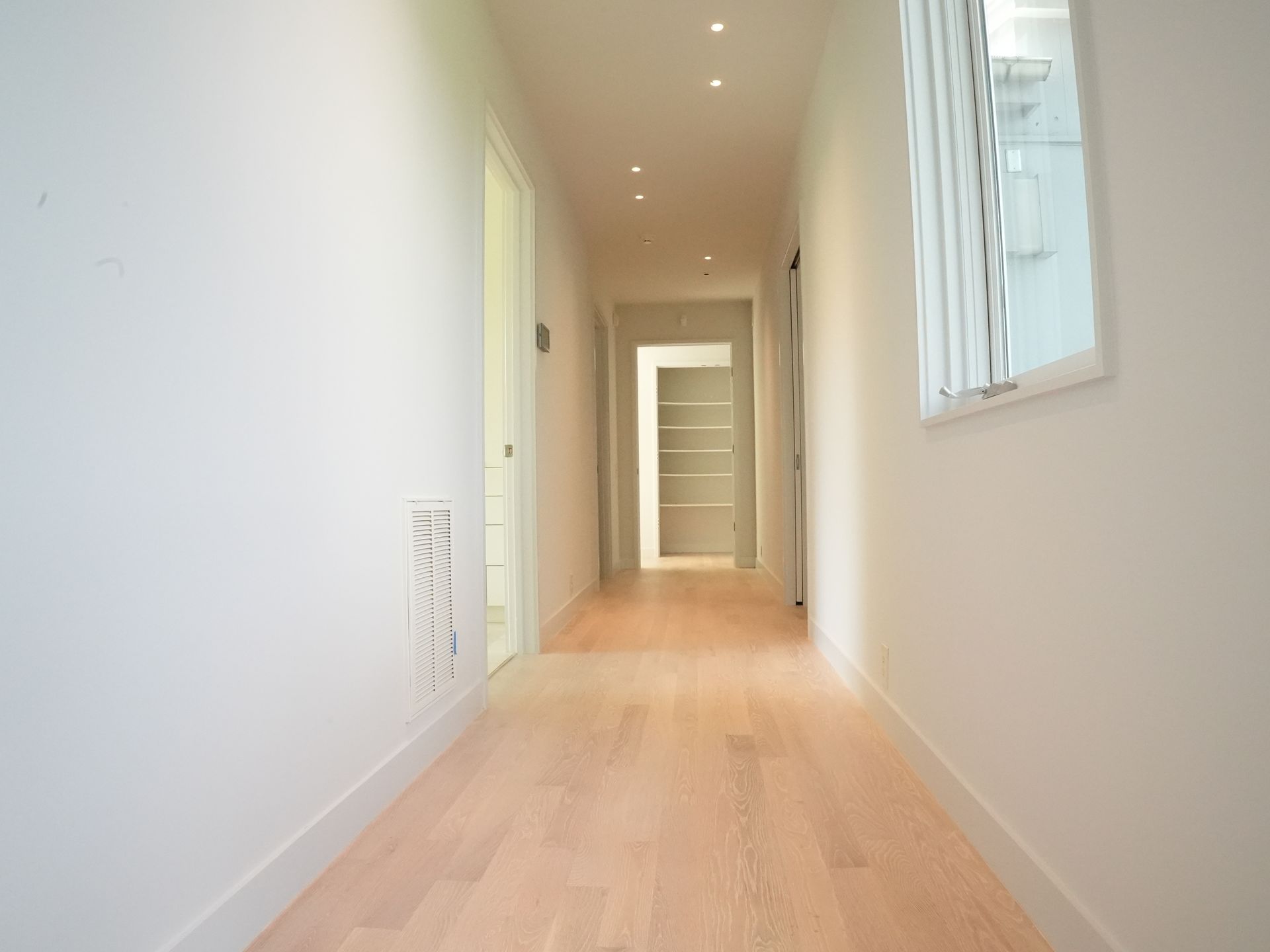 Narrow, white hallway with white oak wood floor, recessed lights, and small window.