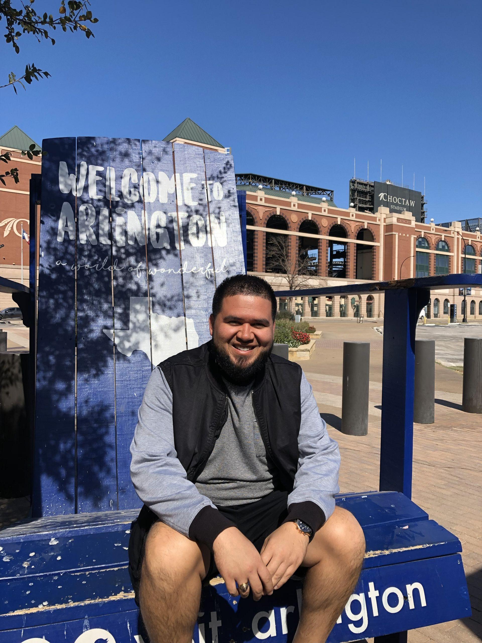Person smiling, sitting on a blue "Welcome to Arlington" bench; background is a baseball stadium under a blue sky.