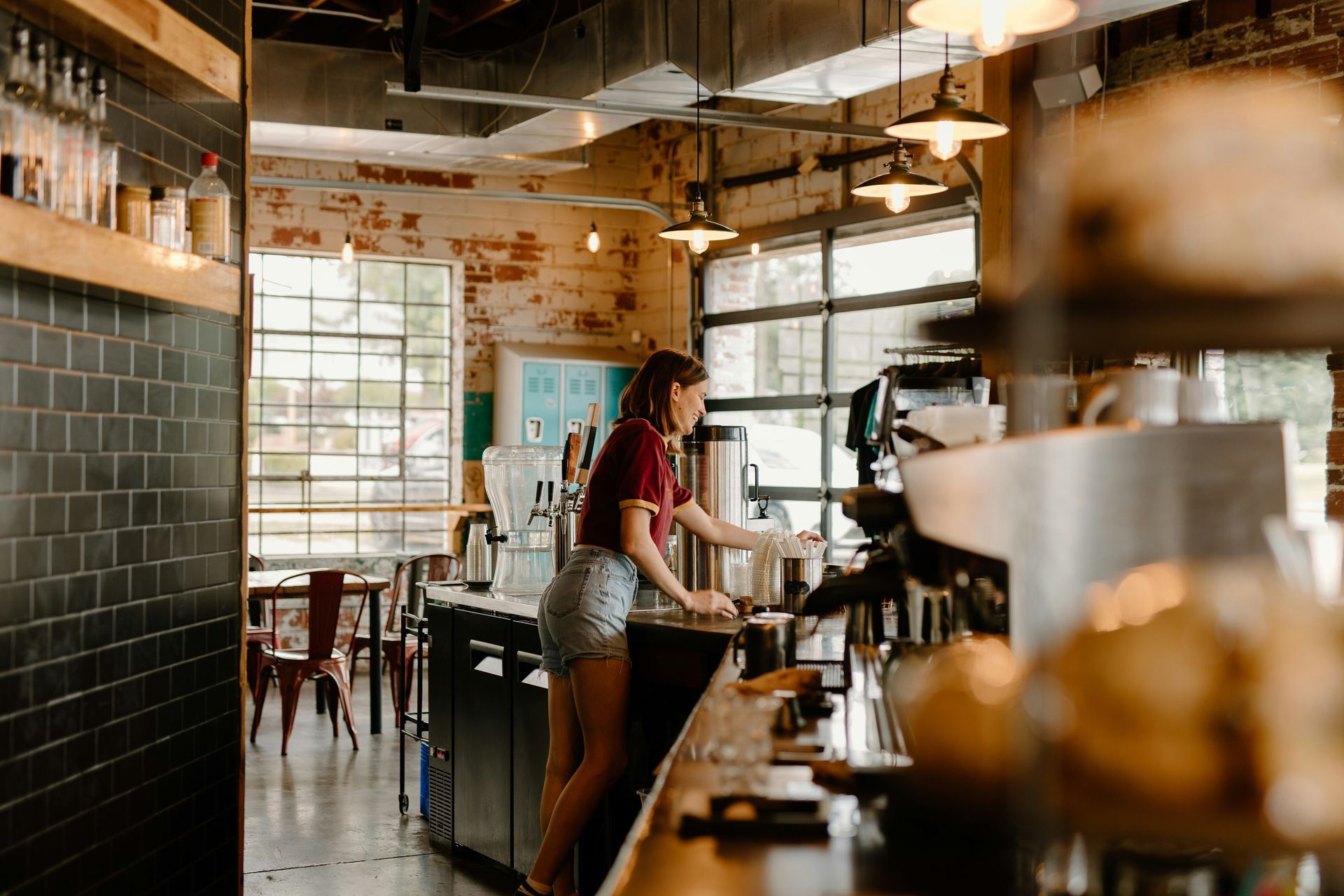 Woman at coffee shop counter preparing drinks. Brick wall, sunlight, industrial interior.