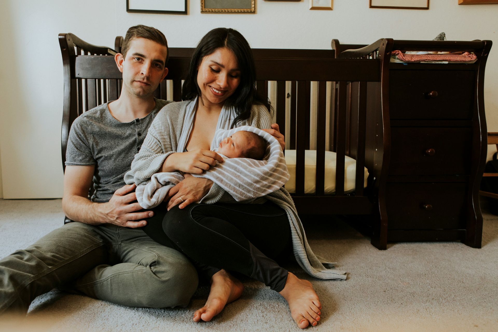 Parents sit on floor next to crib, mother breastfeeding baby.