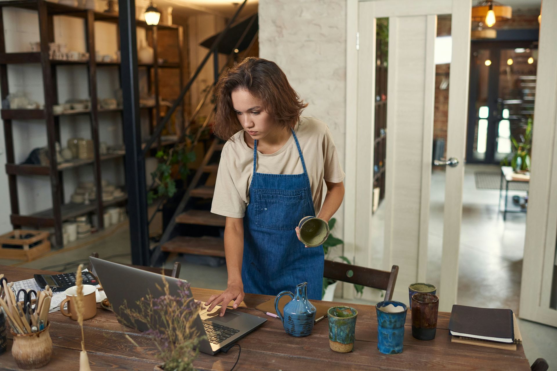 Woman in apron using laptop, examining pottery in a workshop.