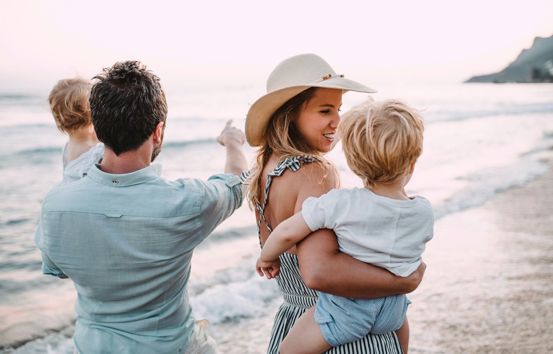 Family on beach, pointing and embracing. Woman with straw hat, holding child. Waves in background.