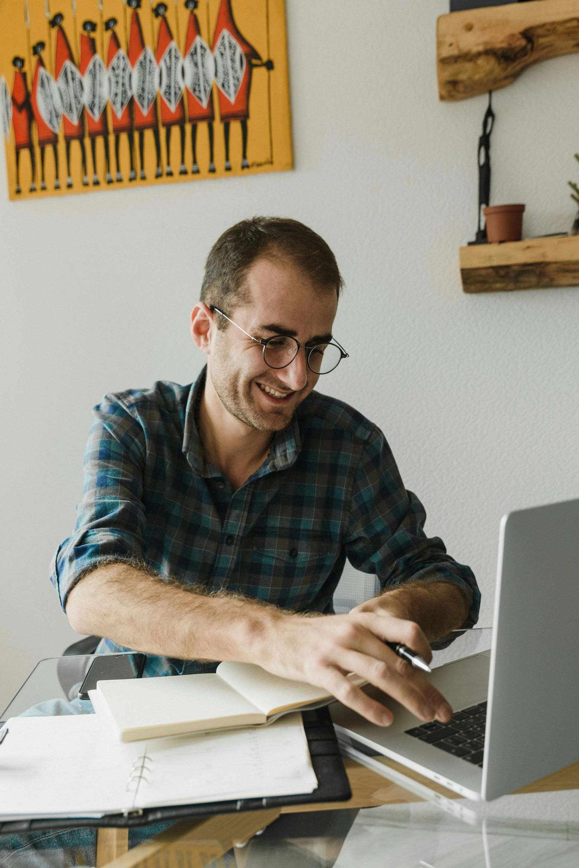 Man in plaid shirt smiles at laptop while working at a desk, notebook open.