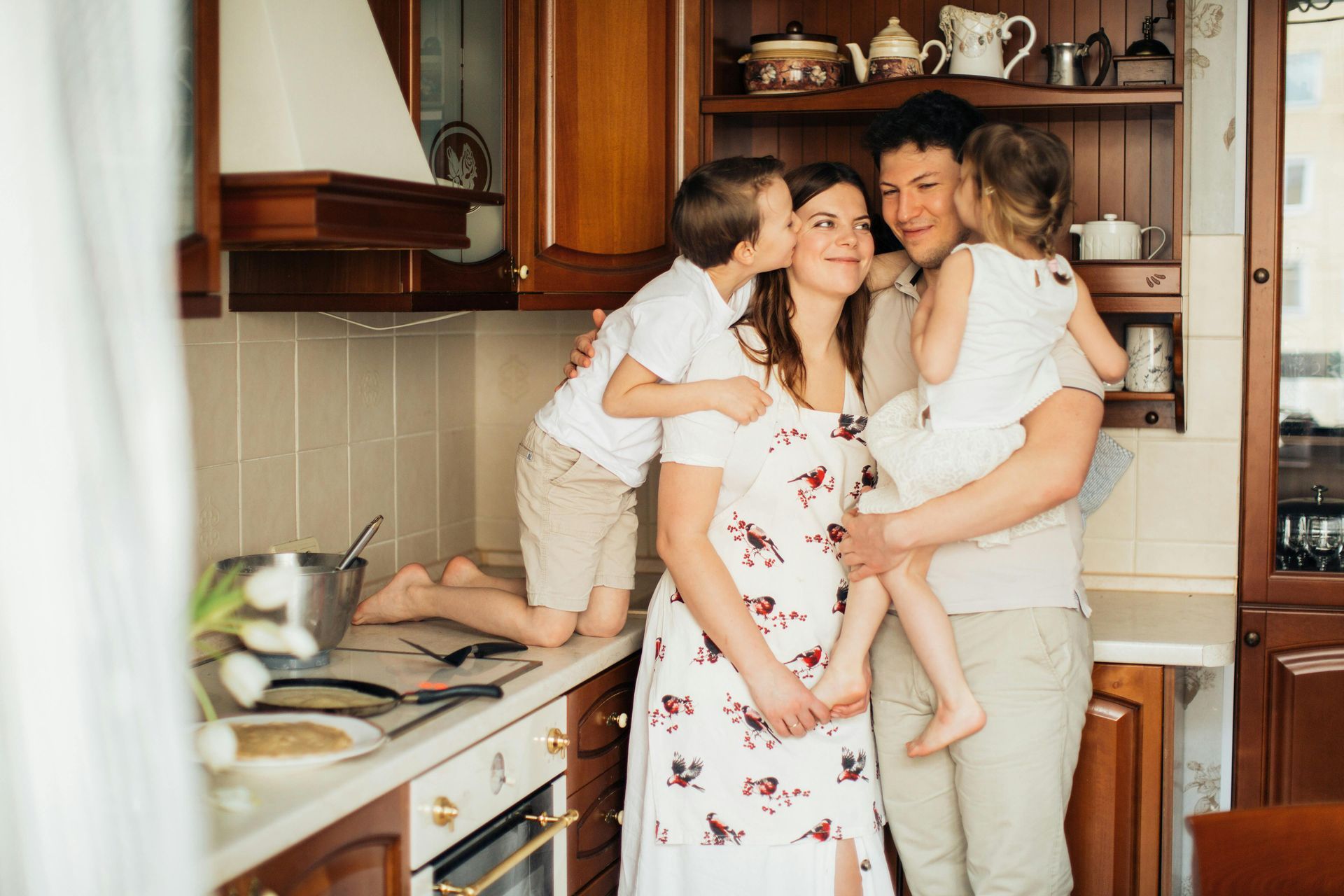 Family in kitchen: Pregnant woman kissed by a boy, held by a man with a girl, near a stove.