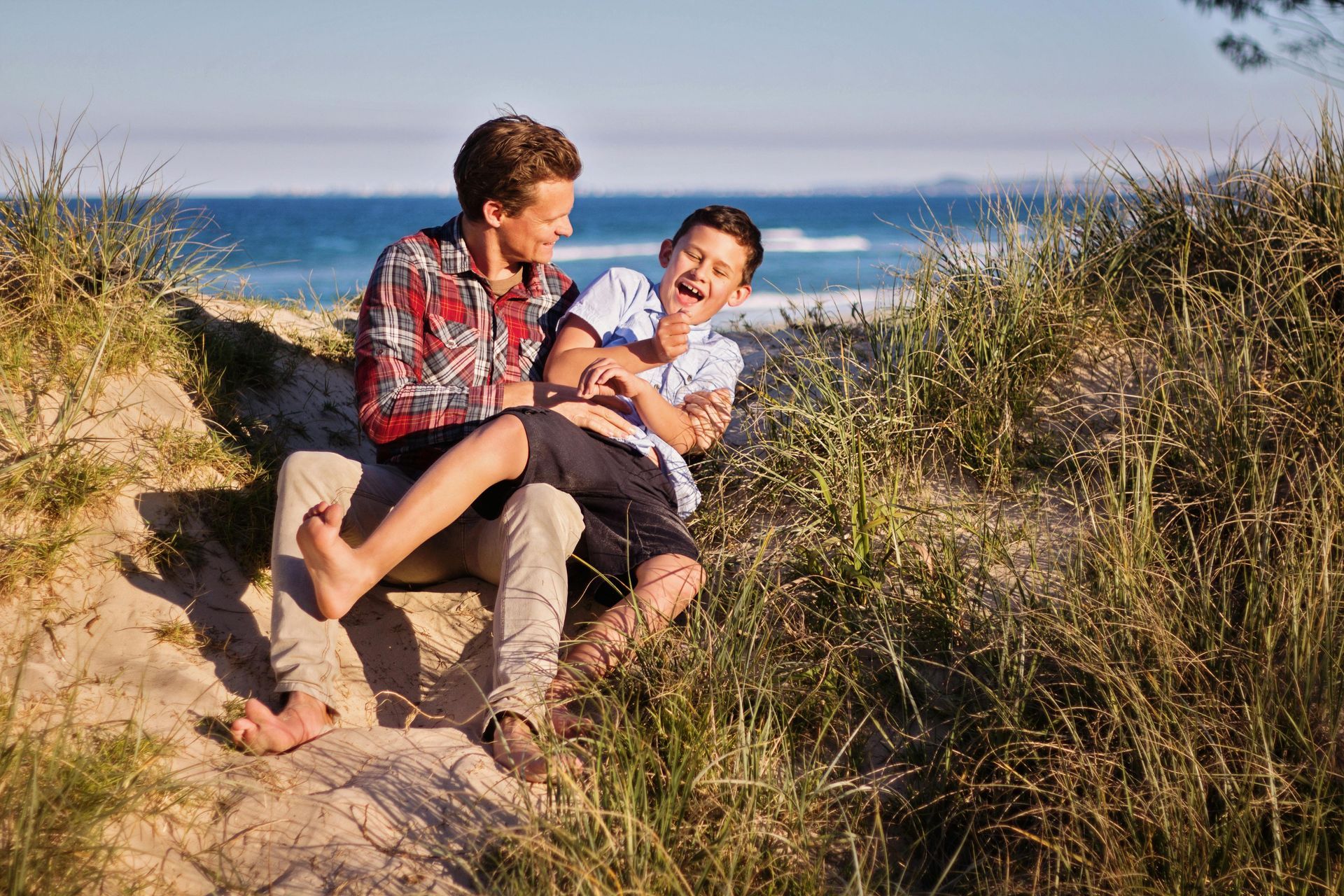 Two people laughing while sitting in beach sand, ocean in the background.