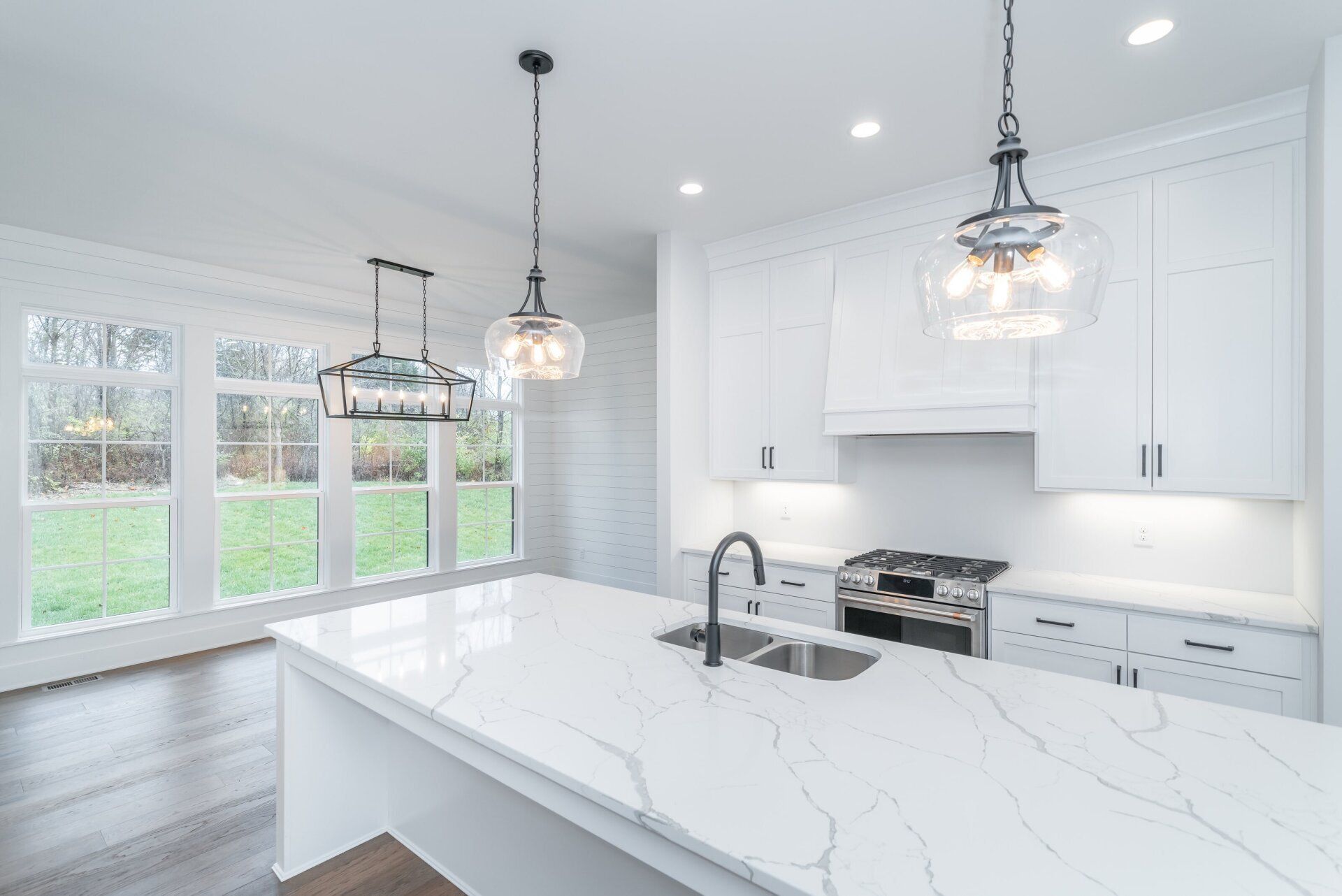 White kitchen with island, range, and hanging lights; large windows overlook green yard.