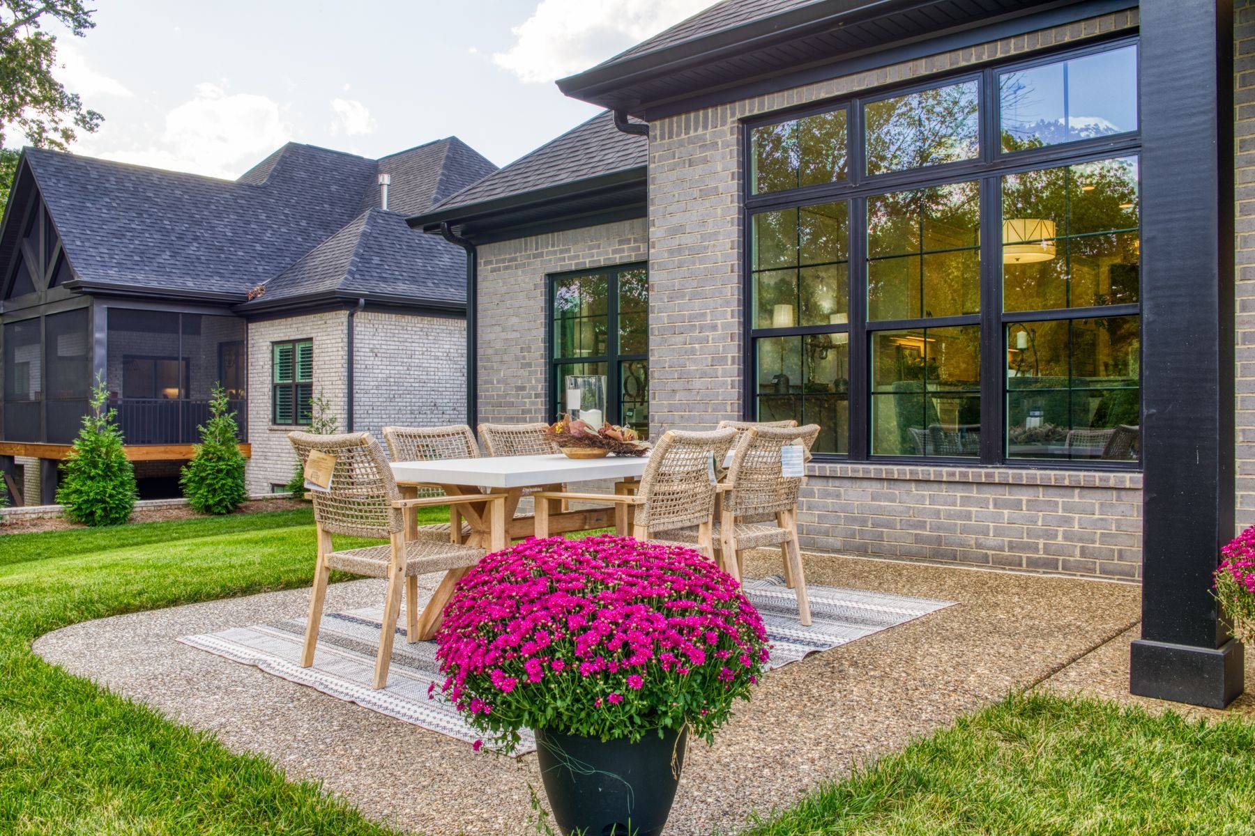 Outdoor patio with table, chairs, and pink flowers in front of a brick house with large windows.