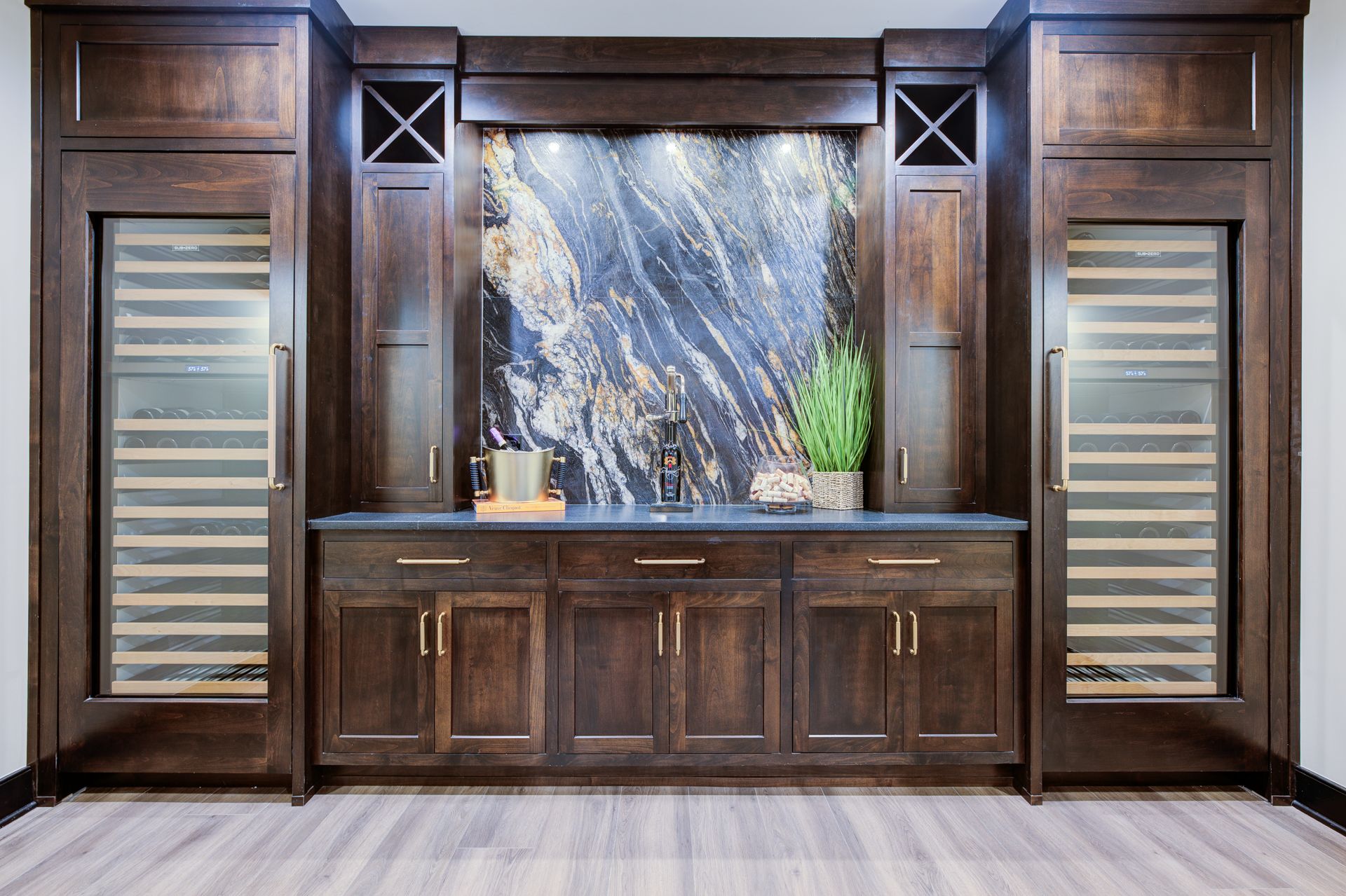 Dark wood built-in bar with wine fridges, marble backsplash, and lower cabinets; small plant on counter.