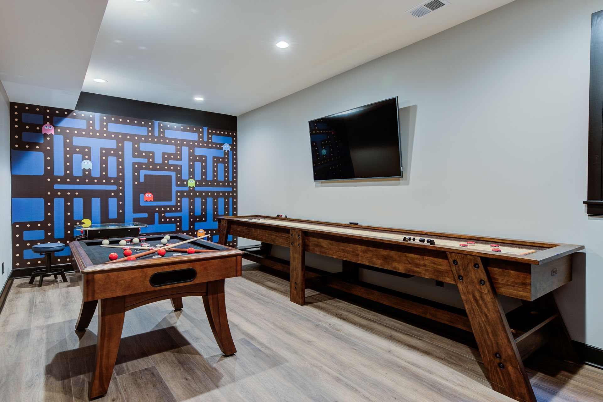 Game room with Pac-Man mural, foosball table, shuffleboard table, and TV on the wall.