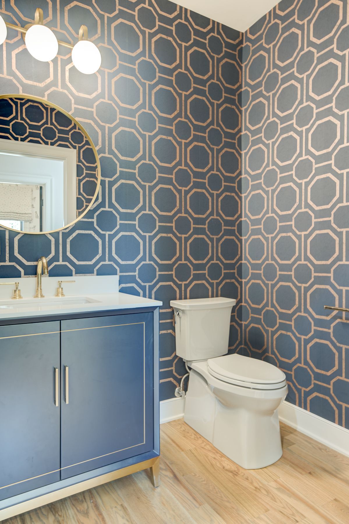 Blue bathroom with geometric patterned wallpaper, vanity, toilet, and round mirror.