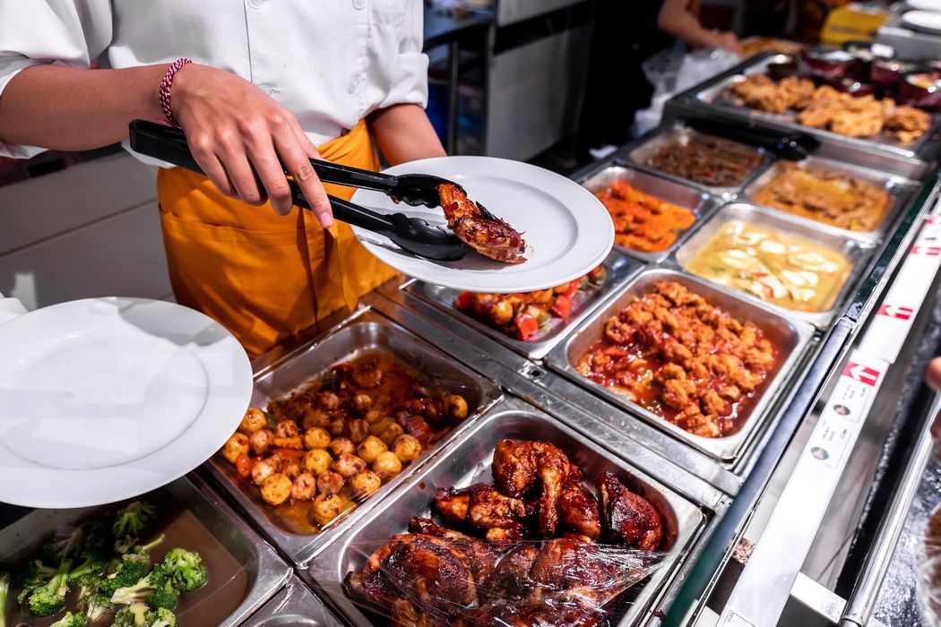 Una persona si serve da un buffet con vassoi in acciaio inossidabile contenenti piatti assortiti e piatti bianchi.