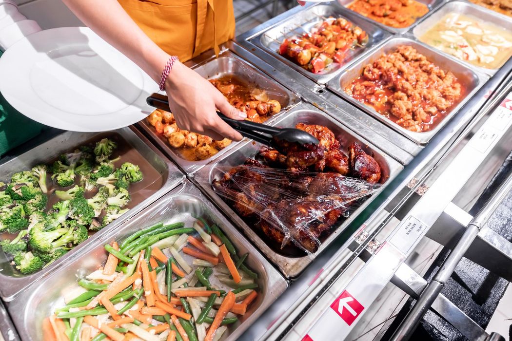 Linea di buffet con vassoi di verdure e carne glassata, con pinze per servire il cibo da un bancone in acciaio inox.