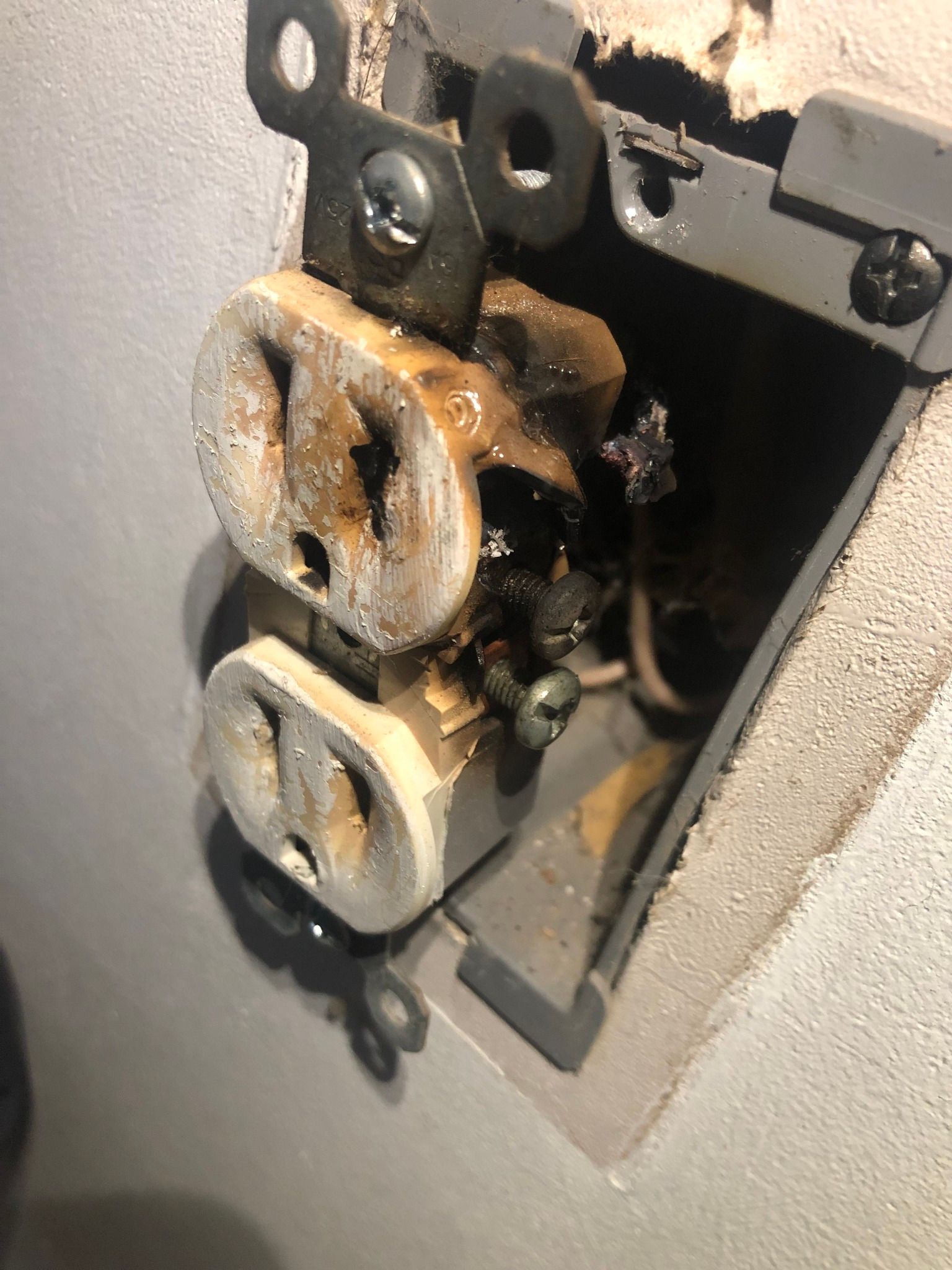 A severely damaged, charred electrical outlet with melted plastic housing mounted in a wall.