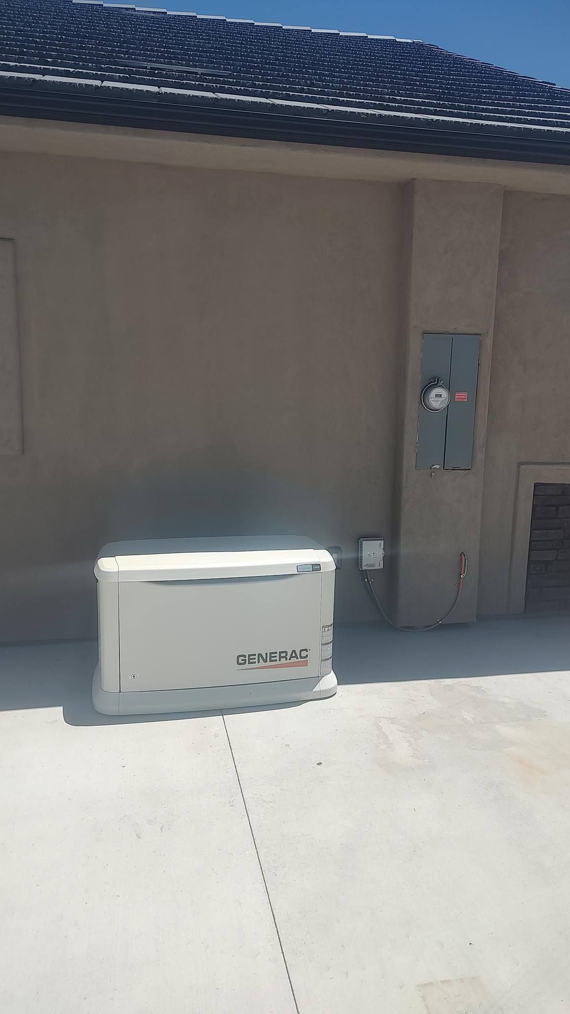 A white Generac home standby generator sits on a concrete pad next to the exterior wall of a tan house.