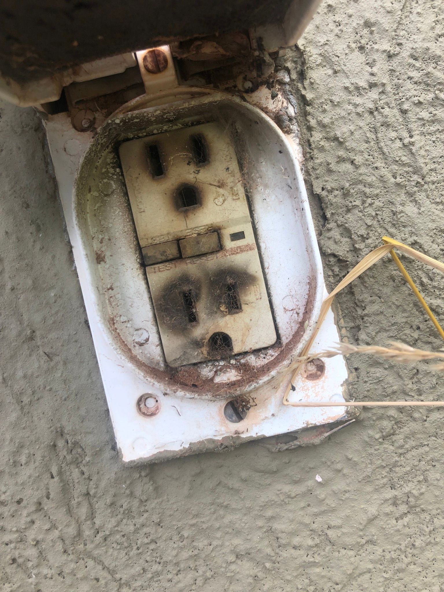 A severely scorched and weathered outdoor electrical outlet mounted on a grey concrete wall.