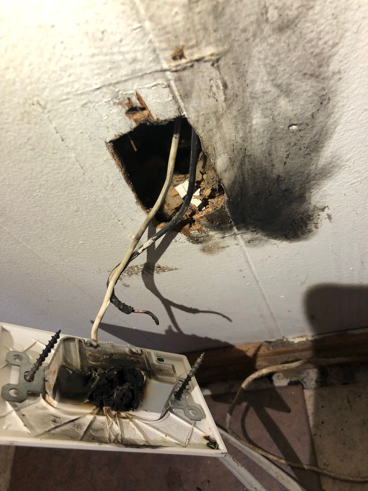 An electrical outlet hanging from a wall, showing significant heat damage, melted plastic, and black soot residue.