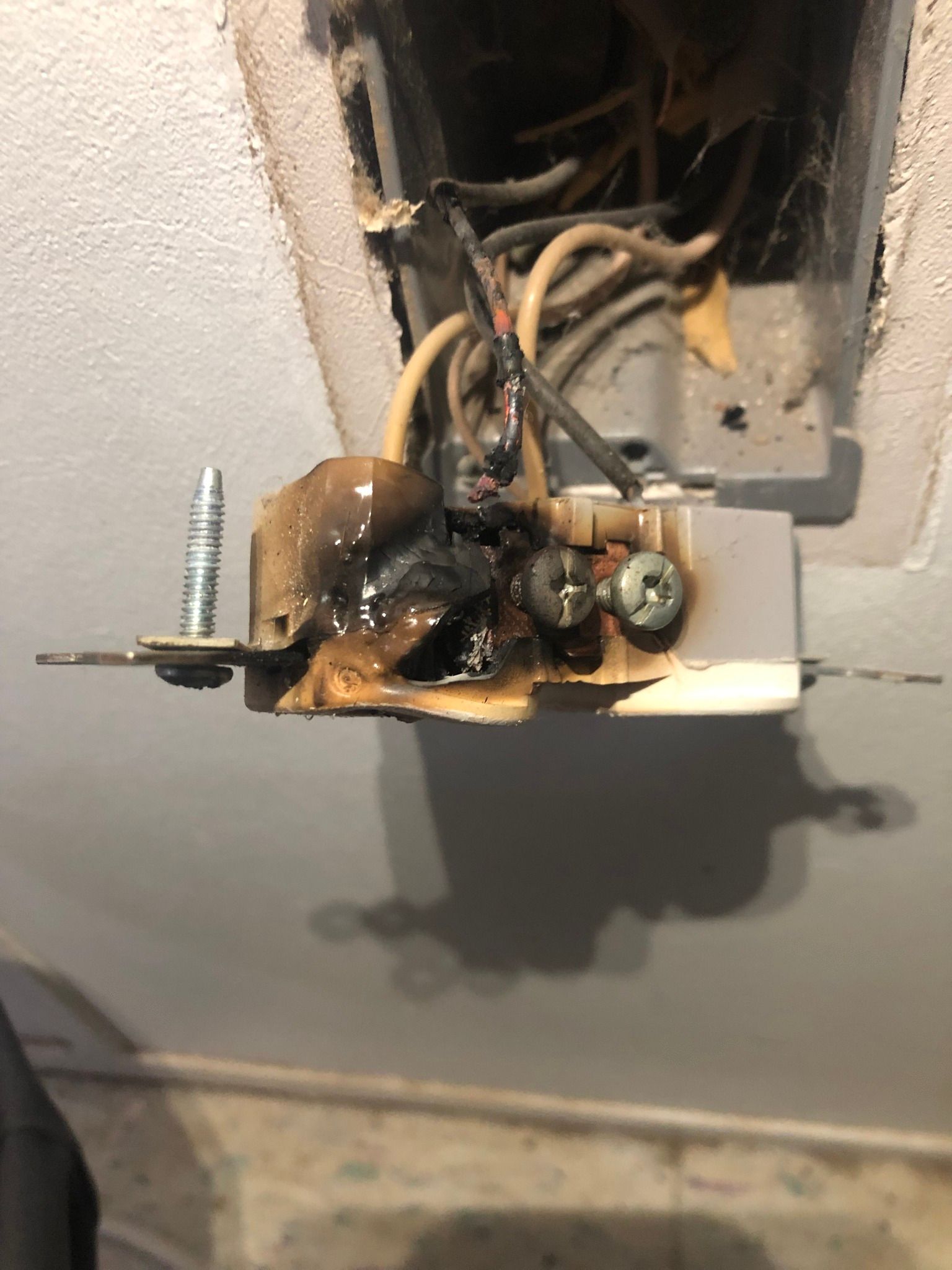 An electrical wall outlet heavily damaged by heat and melting, revealing charred plastic and exposed wiring.