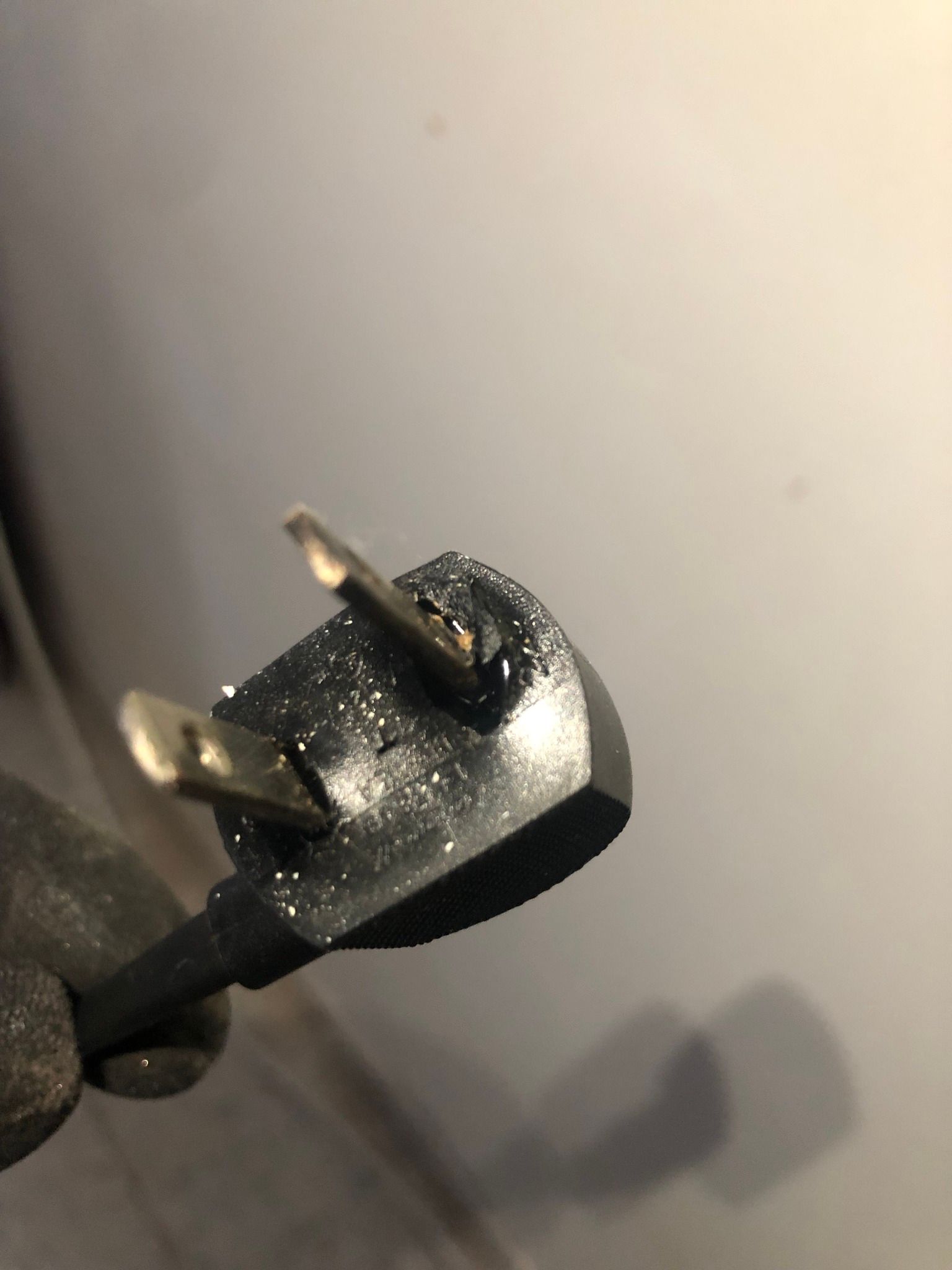 A close-up of a damaged, charred black electrical plug with a warped prong being held by a gloved hand.