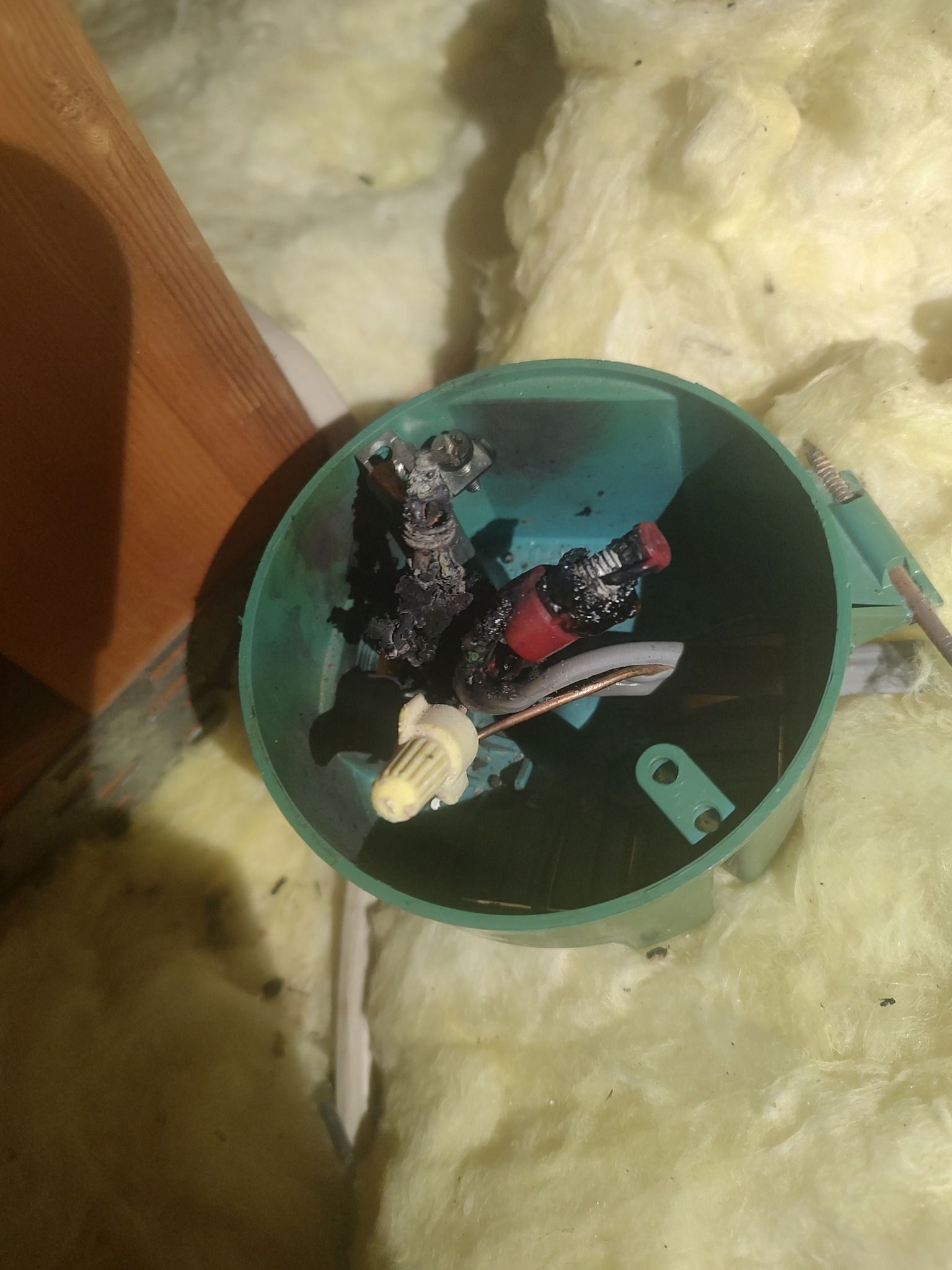 A green electrical box in an attic filled with burnt, charred wires, wire nuts, and insulation debris.