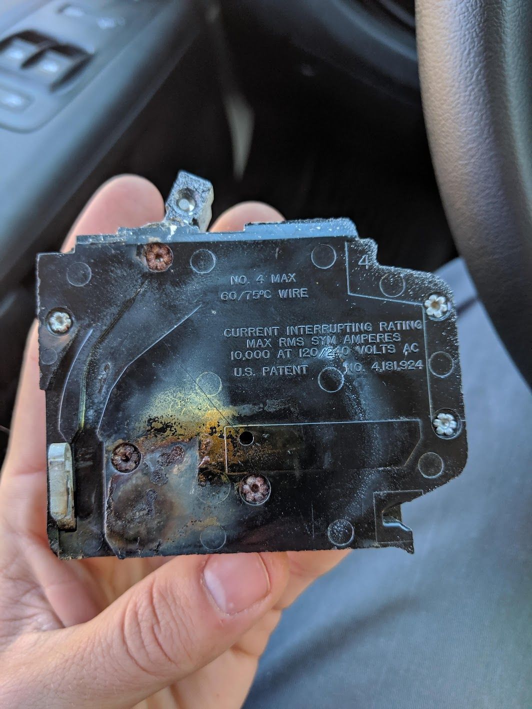 A hand holds a severely charred and melted electrical circuit breaker, showing clear signs of heat damage.