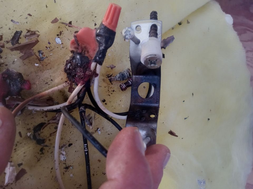 A hand holds a scorched electrical fixture bracket with damaged wiring and a melted orange wire nut against a yellow surface.