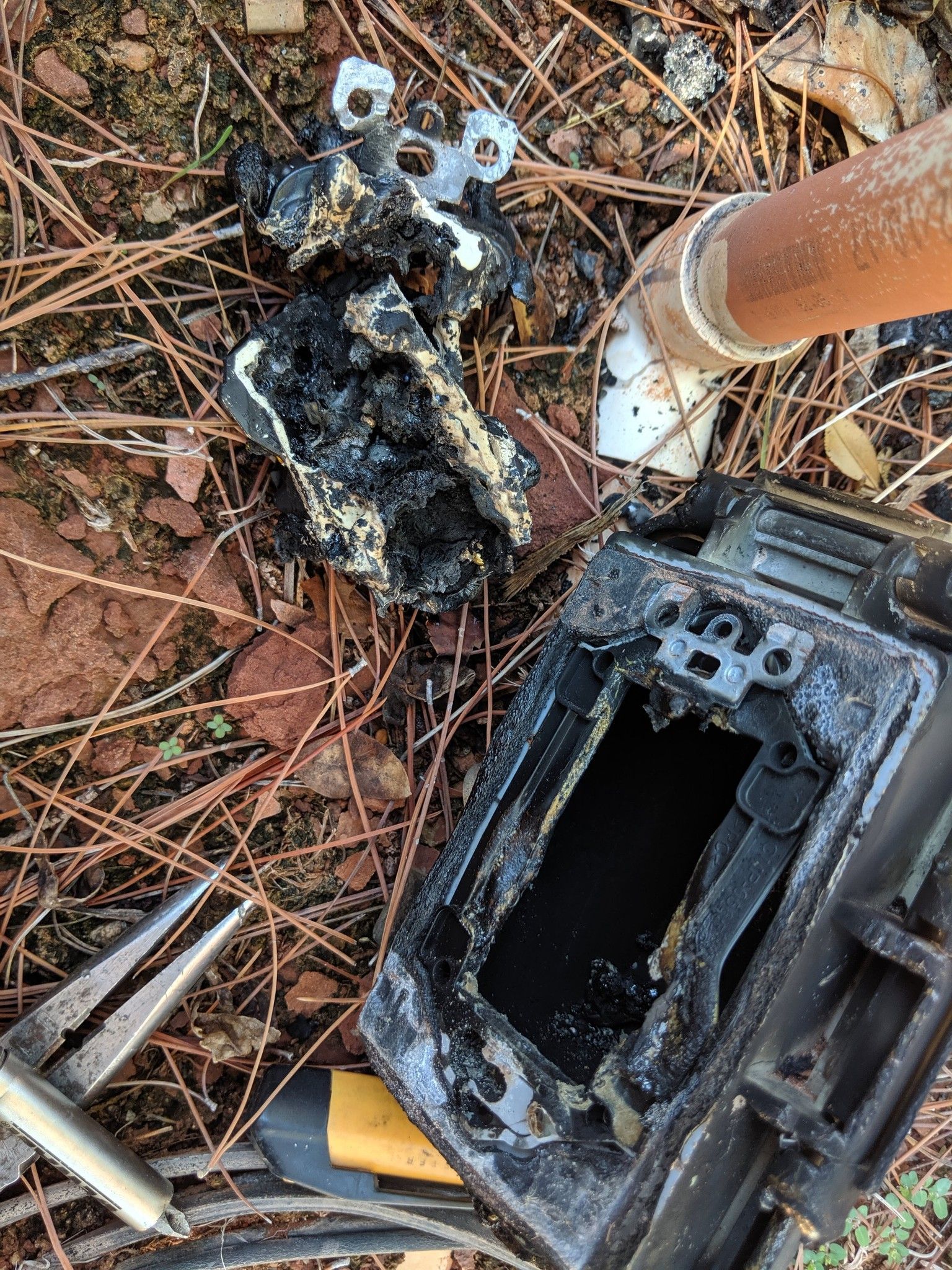 A charred electrical outlet and its plastic housing lying on dry leaves and soil next to a white PVC pipe.