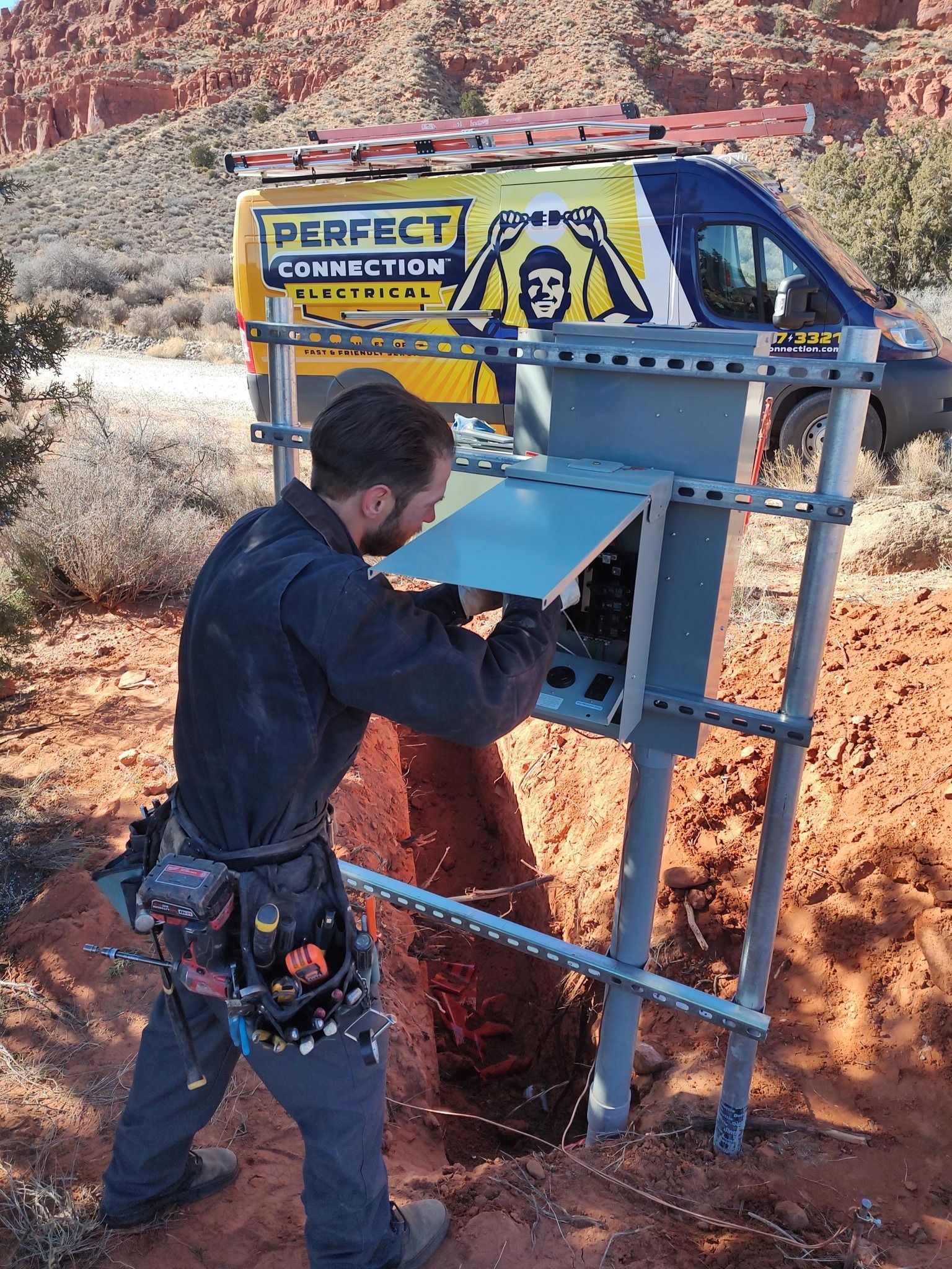 Technician installs electrical equipment on a metal frame at a desert job site with a yellow work van in the background.