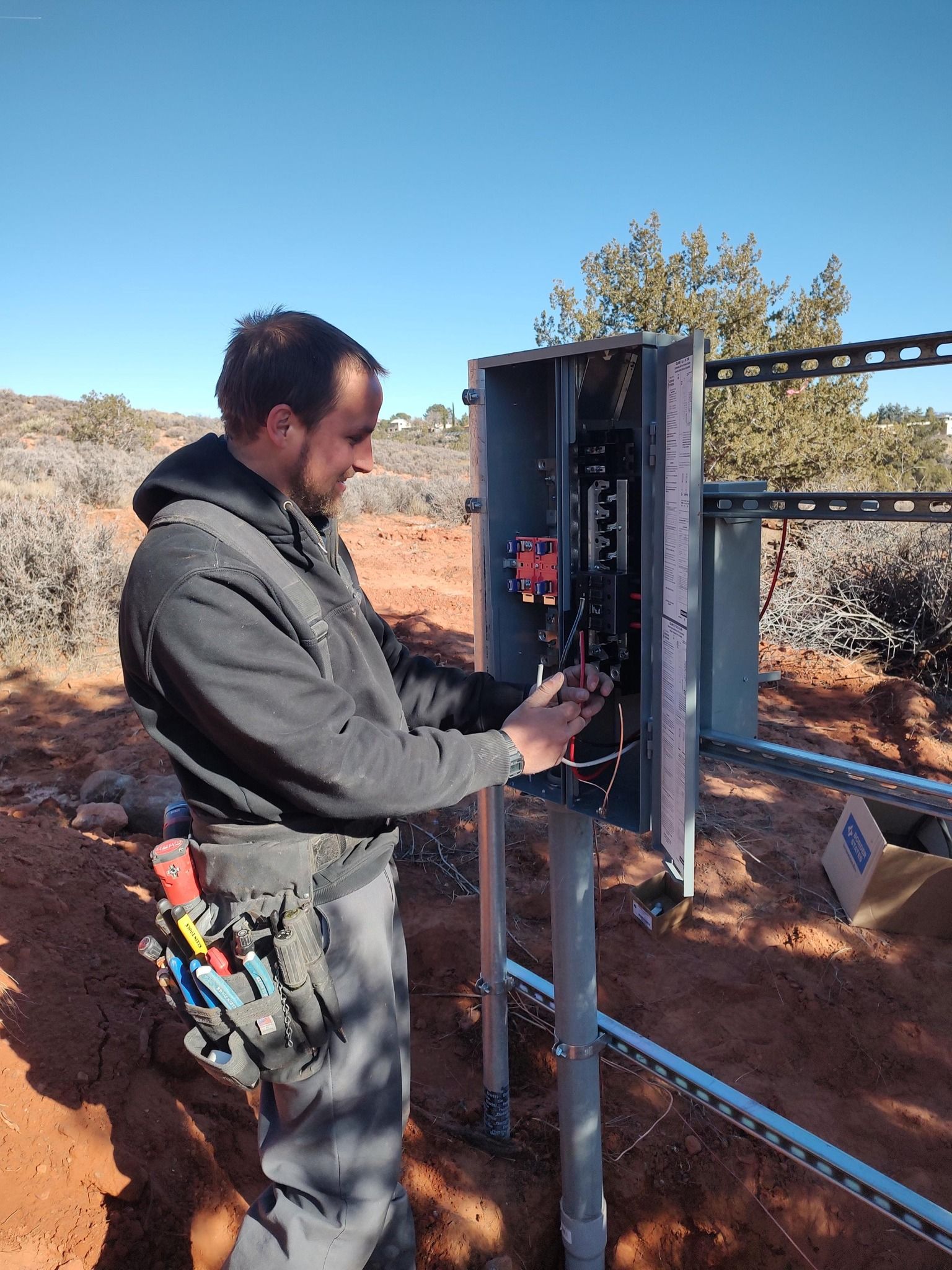 A person works on an electrical panel mounted on a pole in a desert landscape under a clear blue sky.