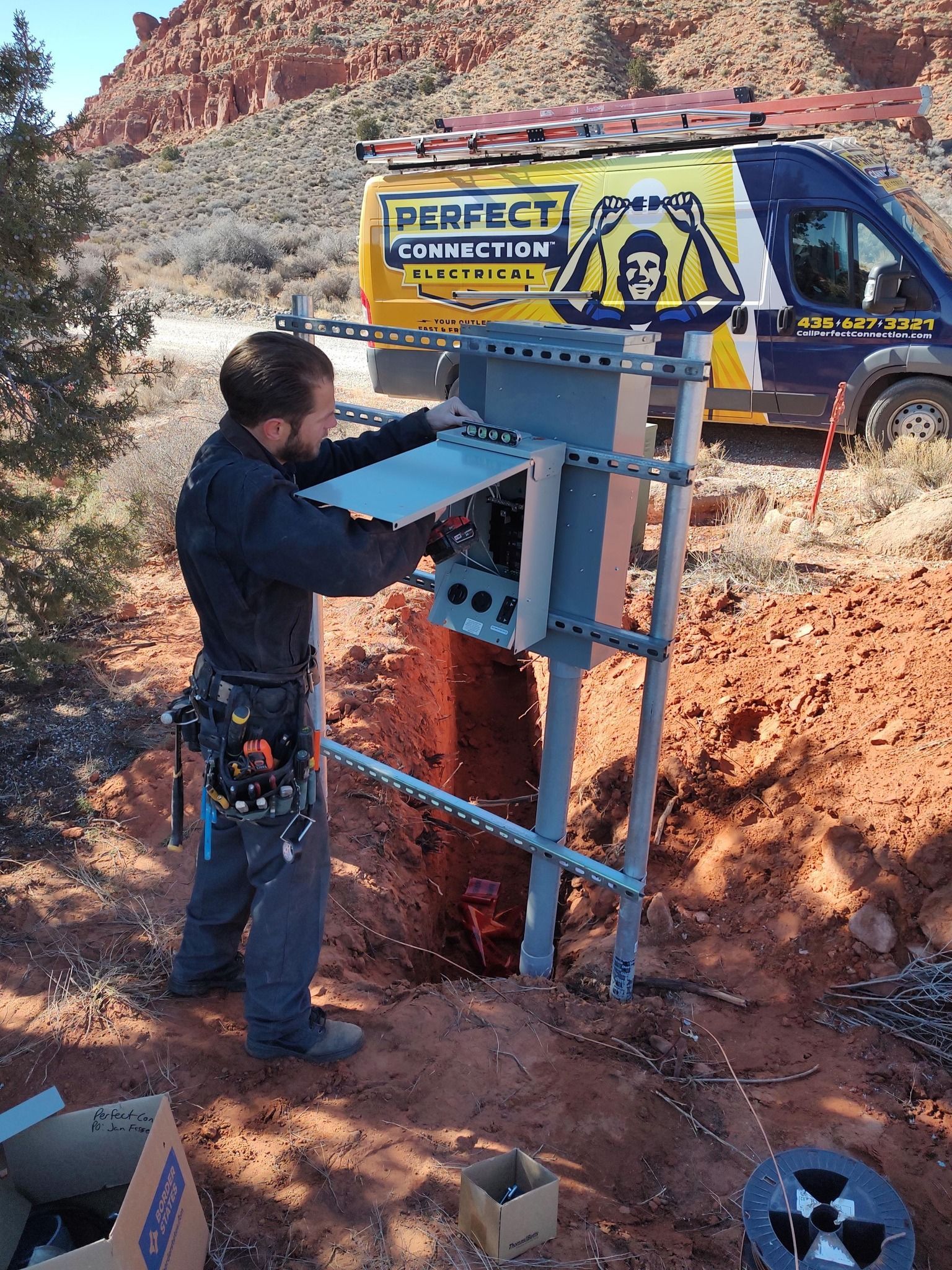 A worker installs electrical equipment on a metal stand at a remote, red-dirt desert construction site near a service van.