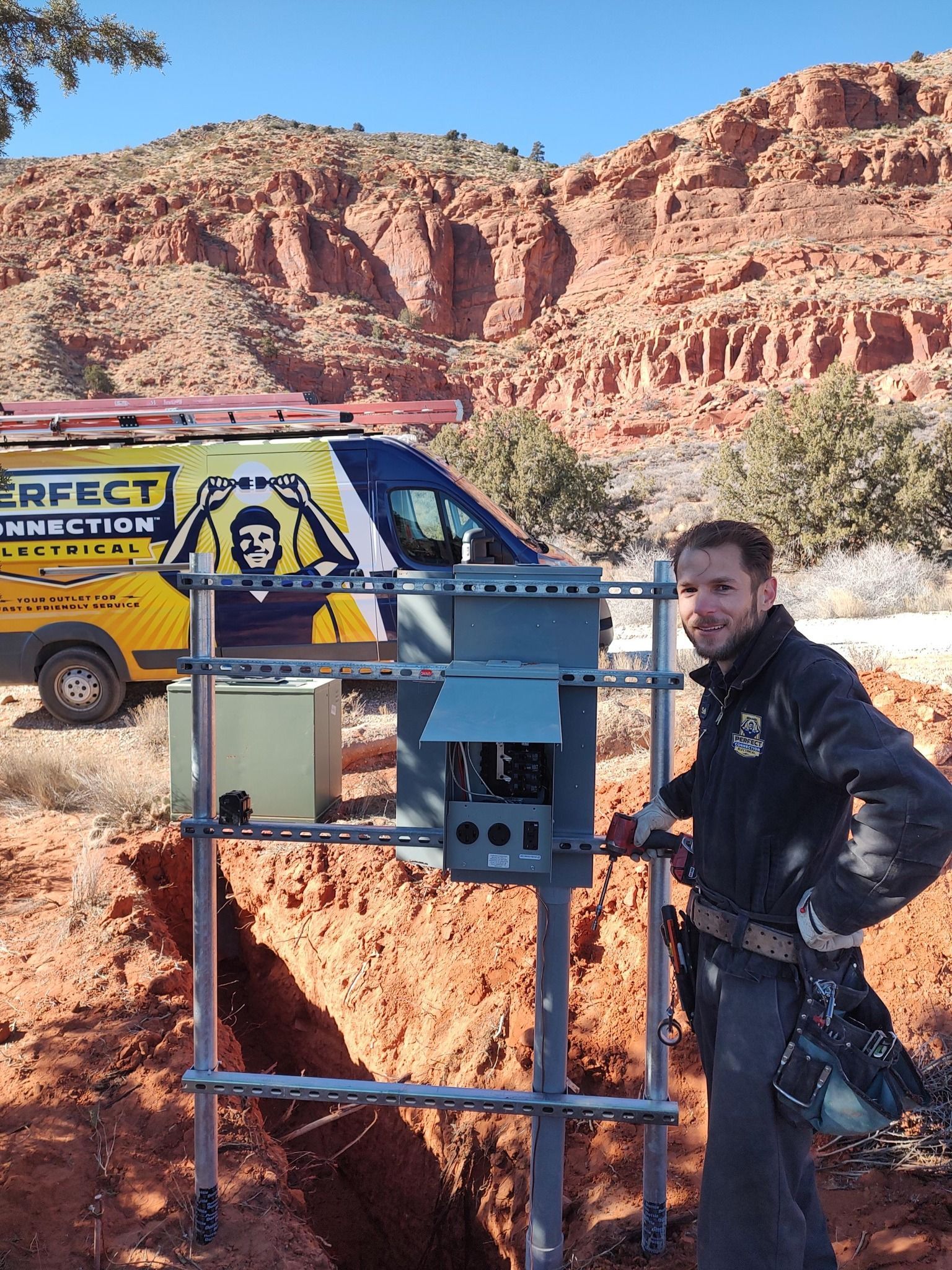 A technician smiles beside an electrical panel mounted on a metal frame above a trench, with a service van nearby.