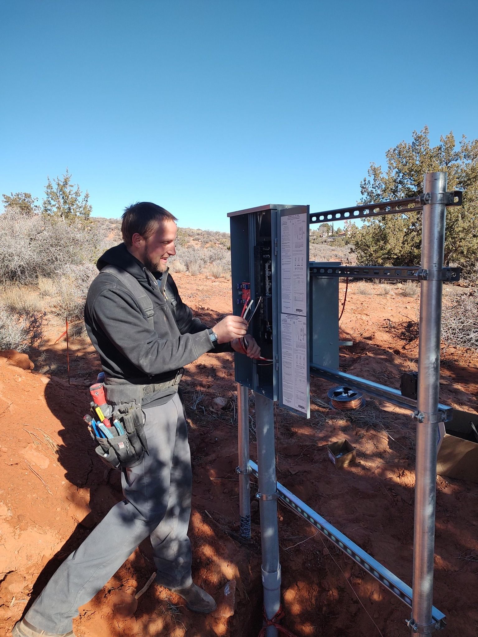 A worker in a dark jacket and tool belt installs electrical equipment on a metal rack frame in a desert landscape.