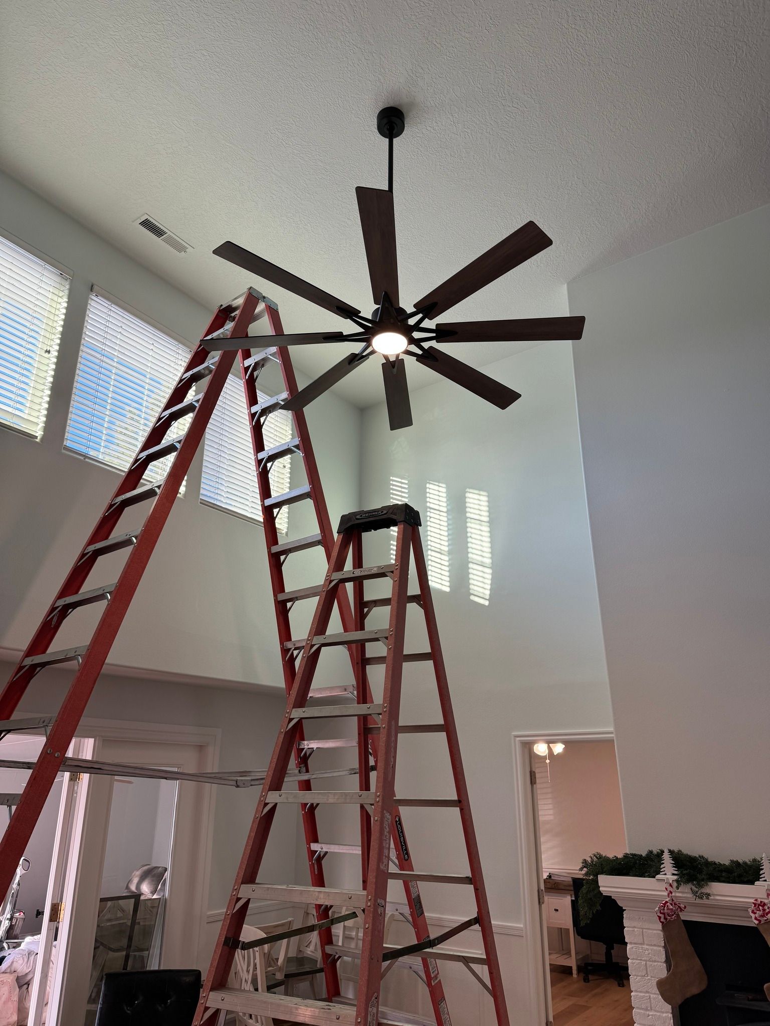 Two tall, red A-frame ladders are set up under a large ceiling fan in a room with high ceilings.