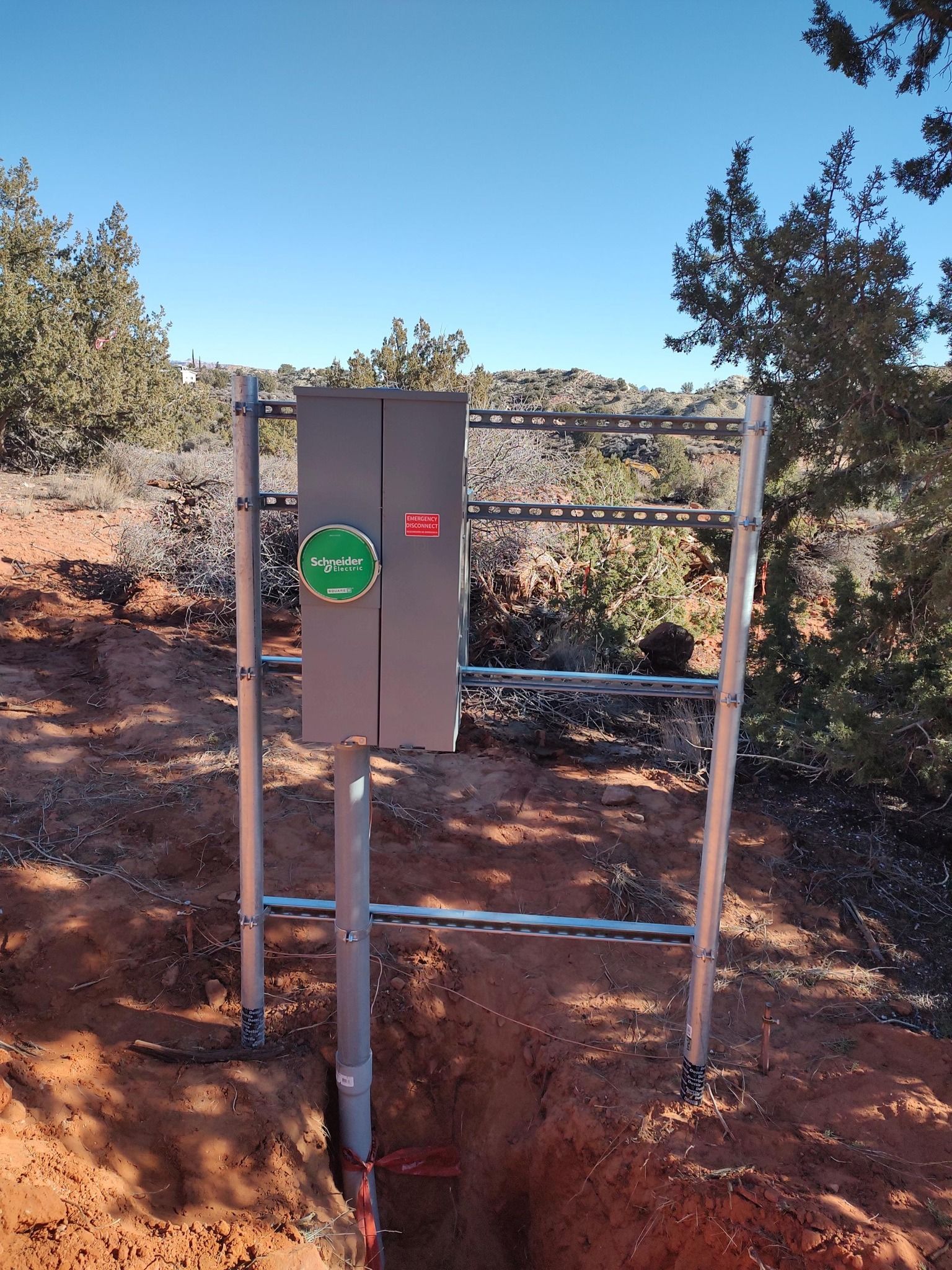 A metal electrical panel mounted on a post frame over an open trench in a desert landscape.