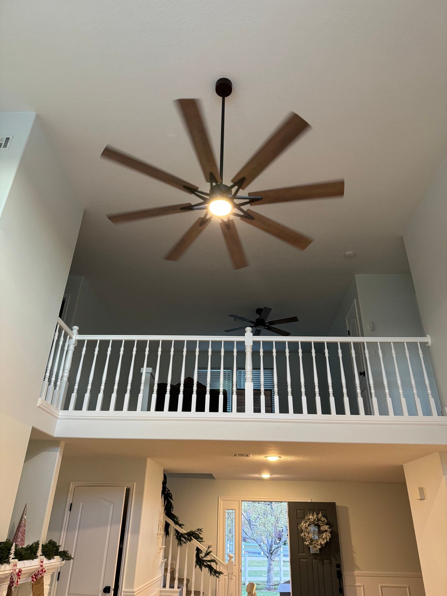 A low-angle view of a vaulted room with an 8-blade ceiling fan, white railing of a loft, and an open front doorway.