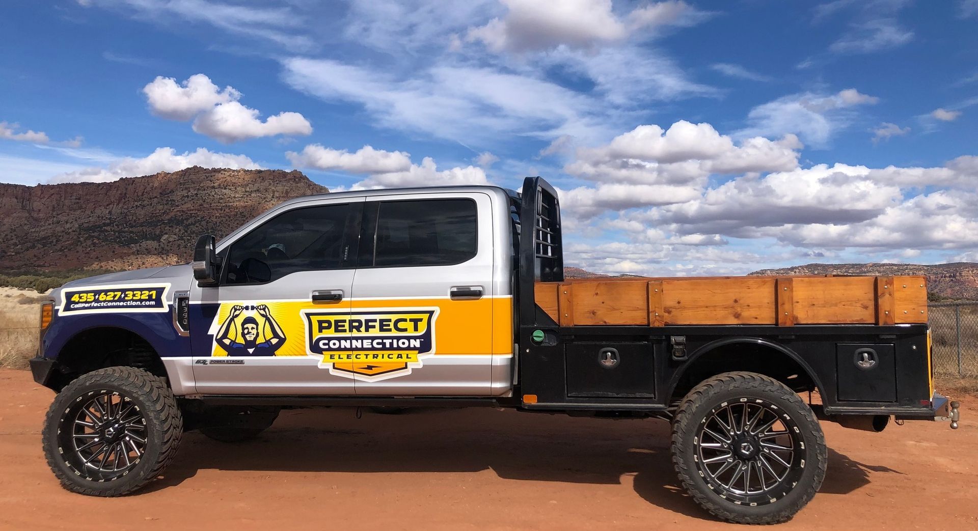 A side view of a customized silver and yellow truck with a wooden flatbed, parked on red dirt against a cloudy sky.
