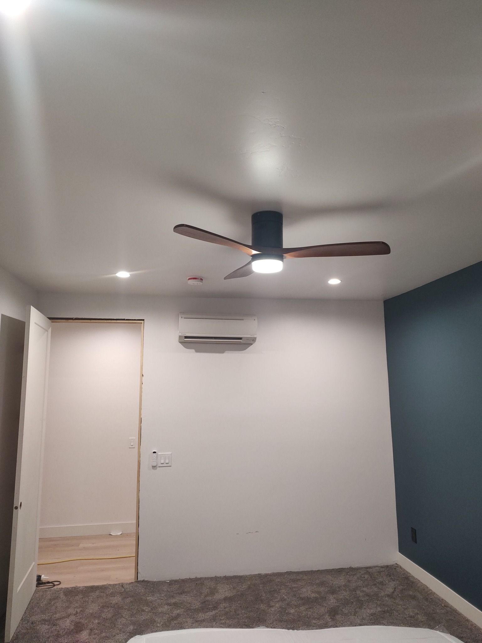 A room with carpeted flooring, a ceiling fan with lights, a wall-mounted mini-split AC, and one dark blue accent wall.