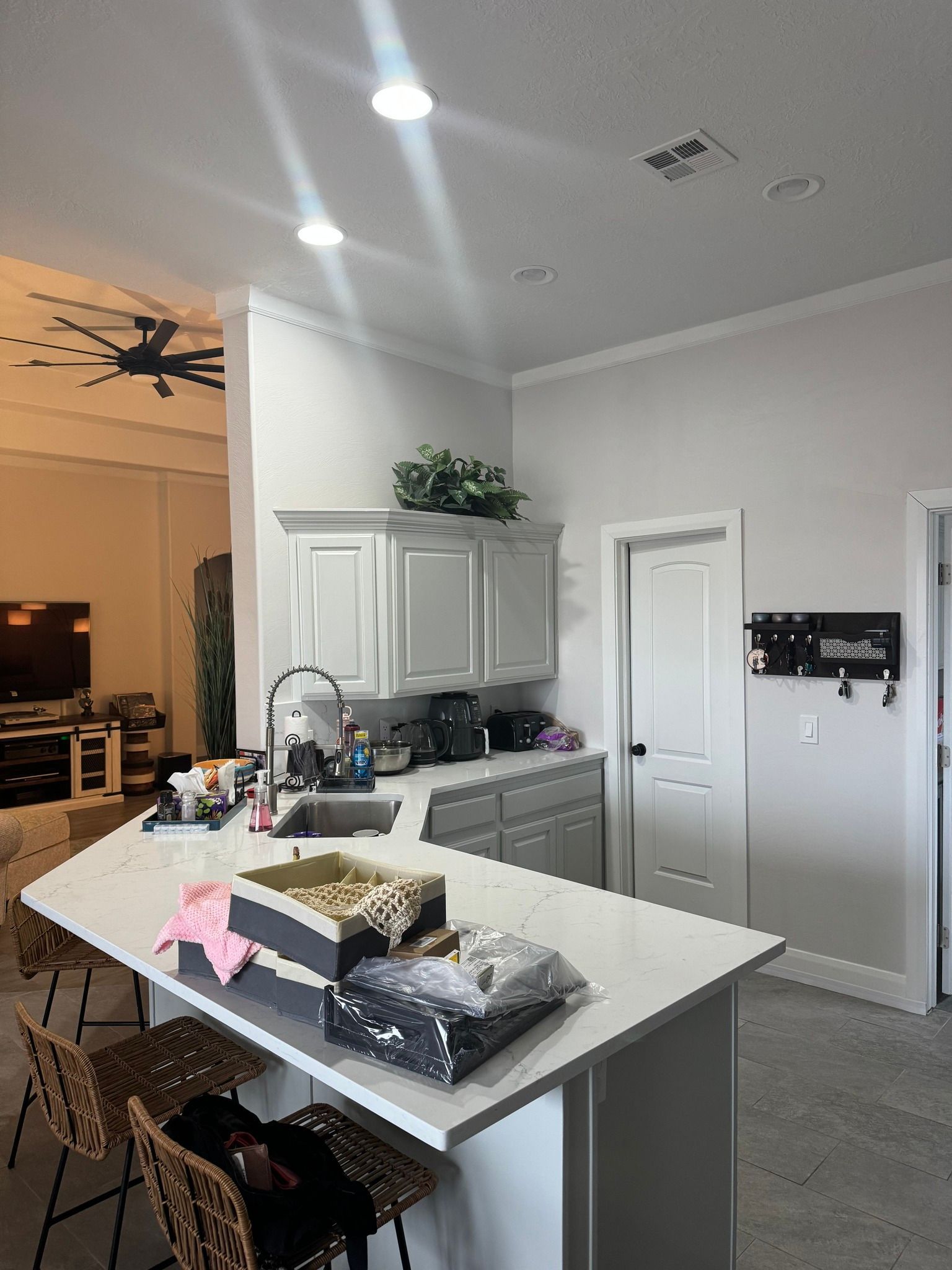 A modern kitchen island with white counters, a sink, and woven stools in a room with light grey walls and recessed lighting.