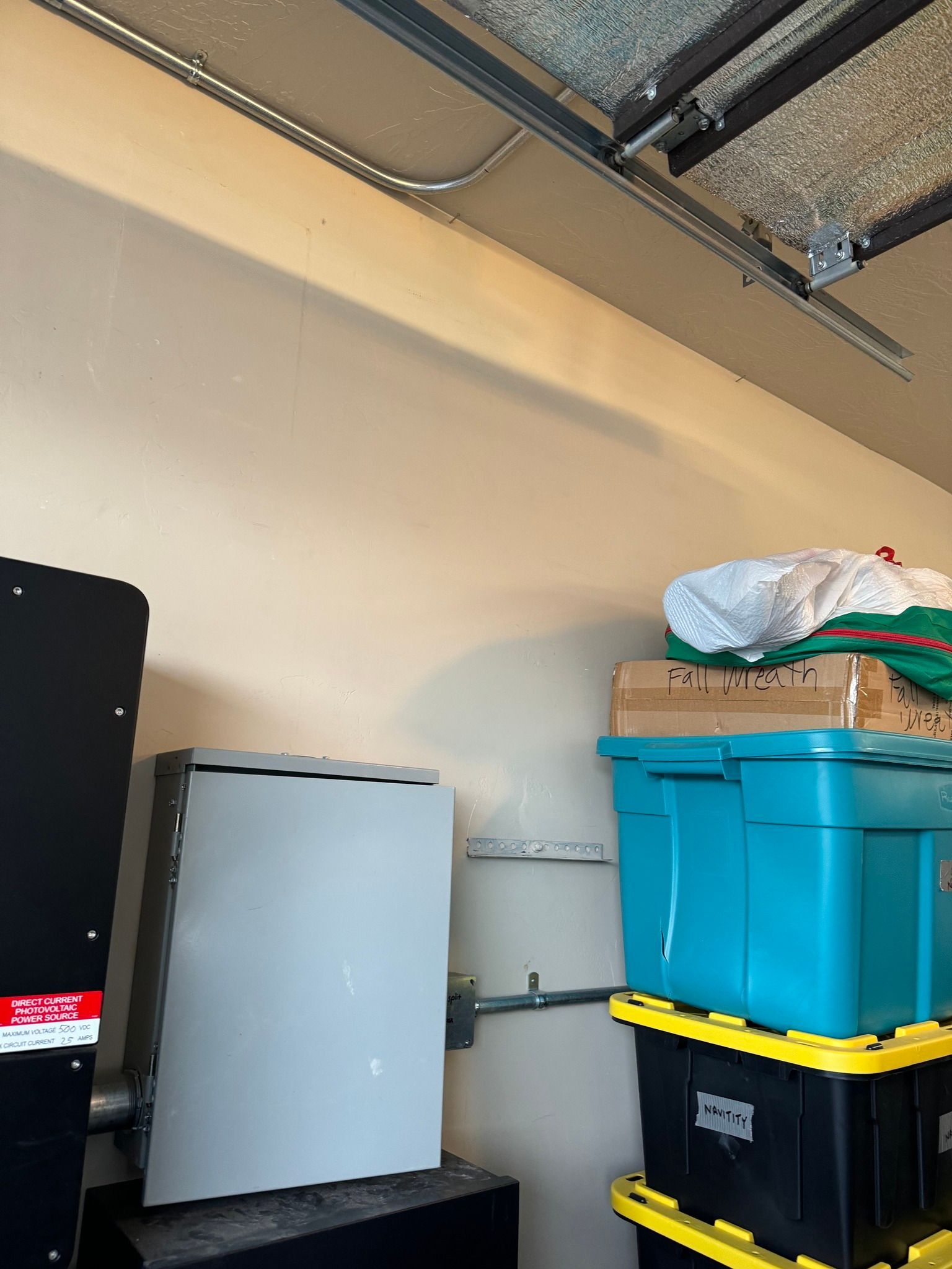 A gray electrical panel mounted on a garage wall, next to stacked storage bins under a ceiling track.