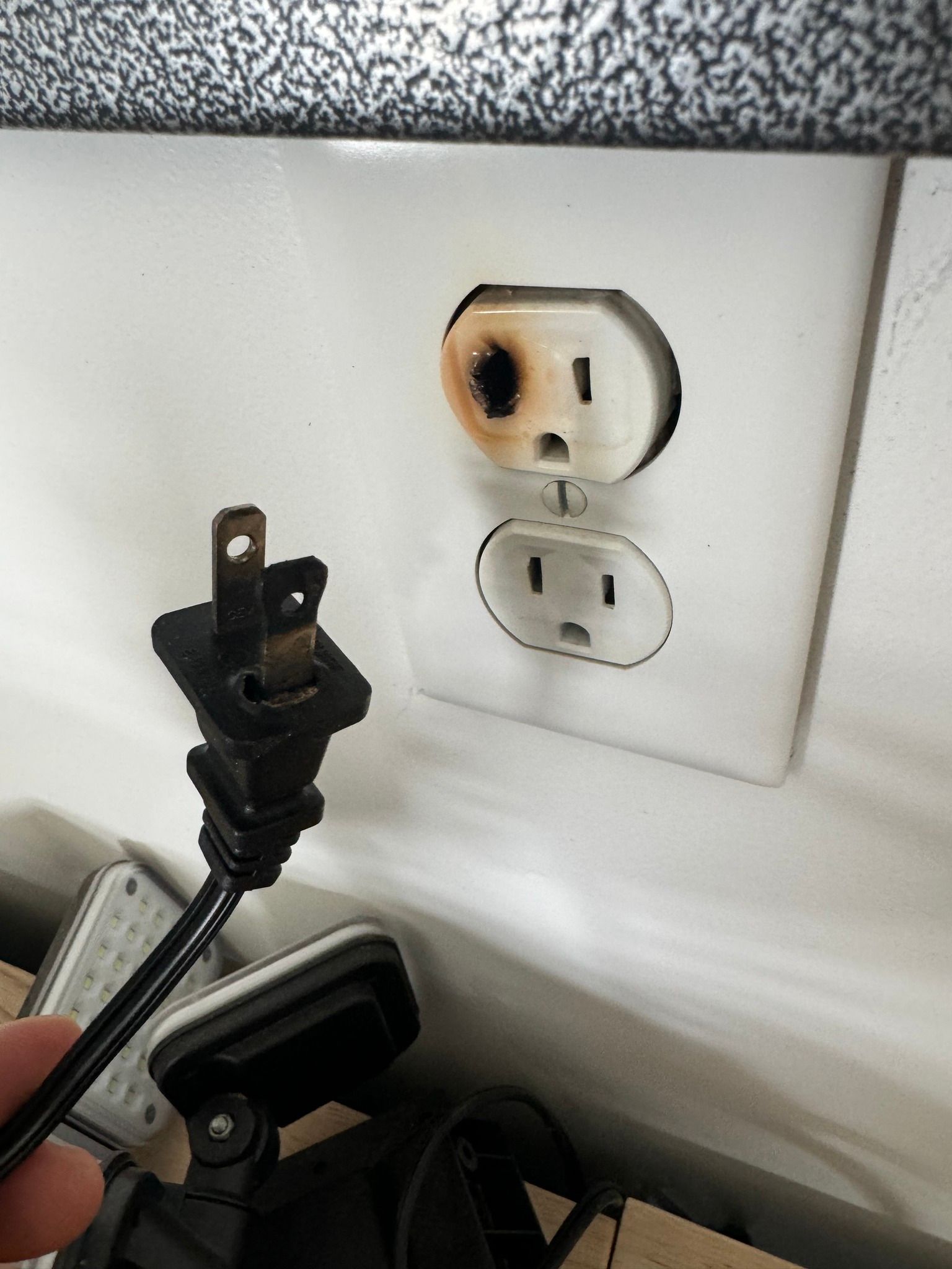 A hand holds a scorched electrical plug next to a wall outlet with visible fire damage around the top socket.