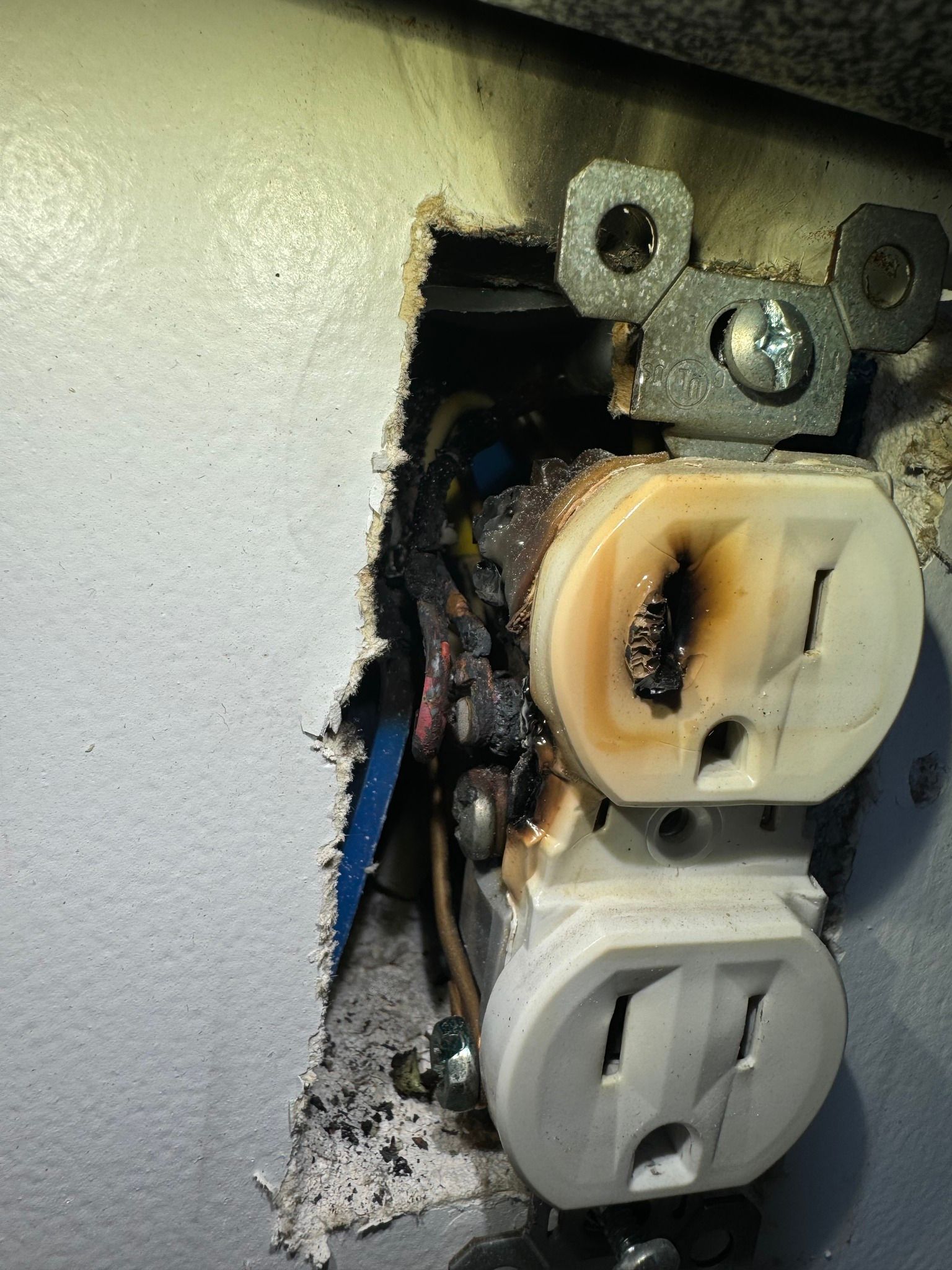 A wall outlet with severe charring and melting damage around the top socket and exposed electrical wiring.