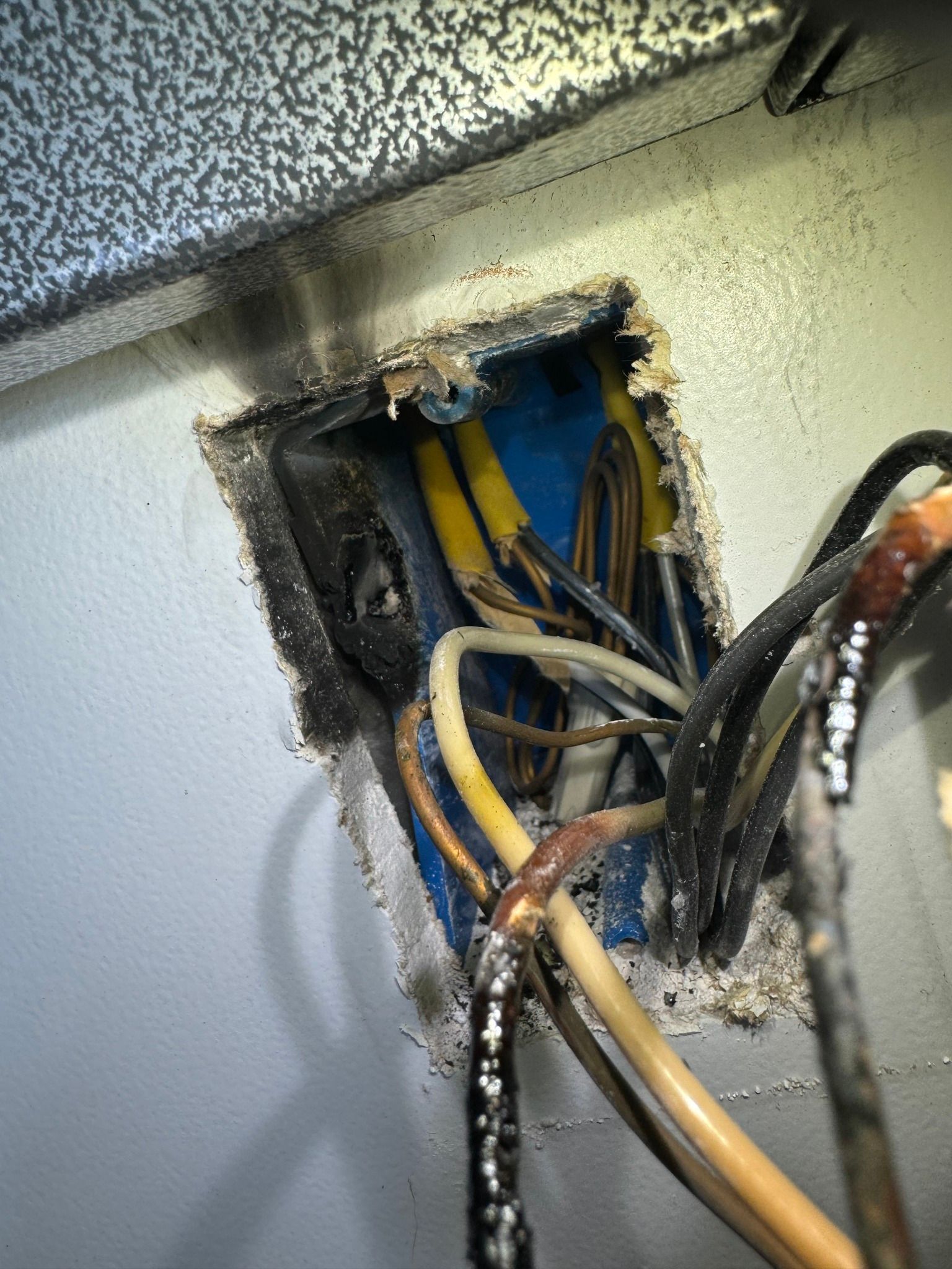 A wall electrical box with scorched wires, melted insulation, and signs of fire damage on the surrounding wall.