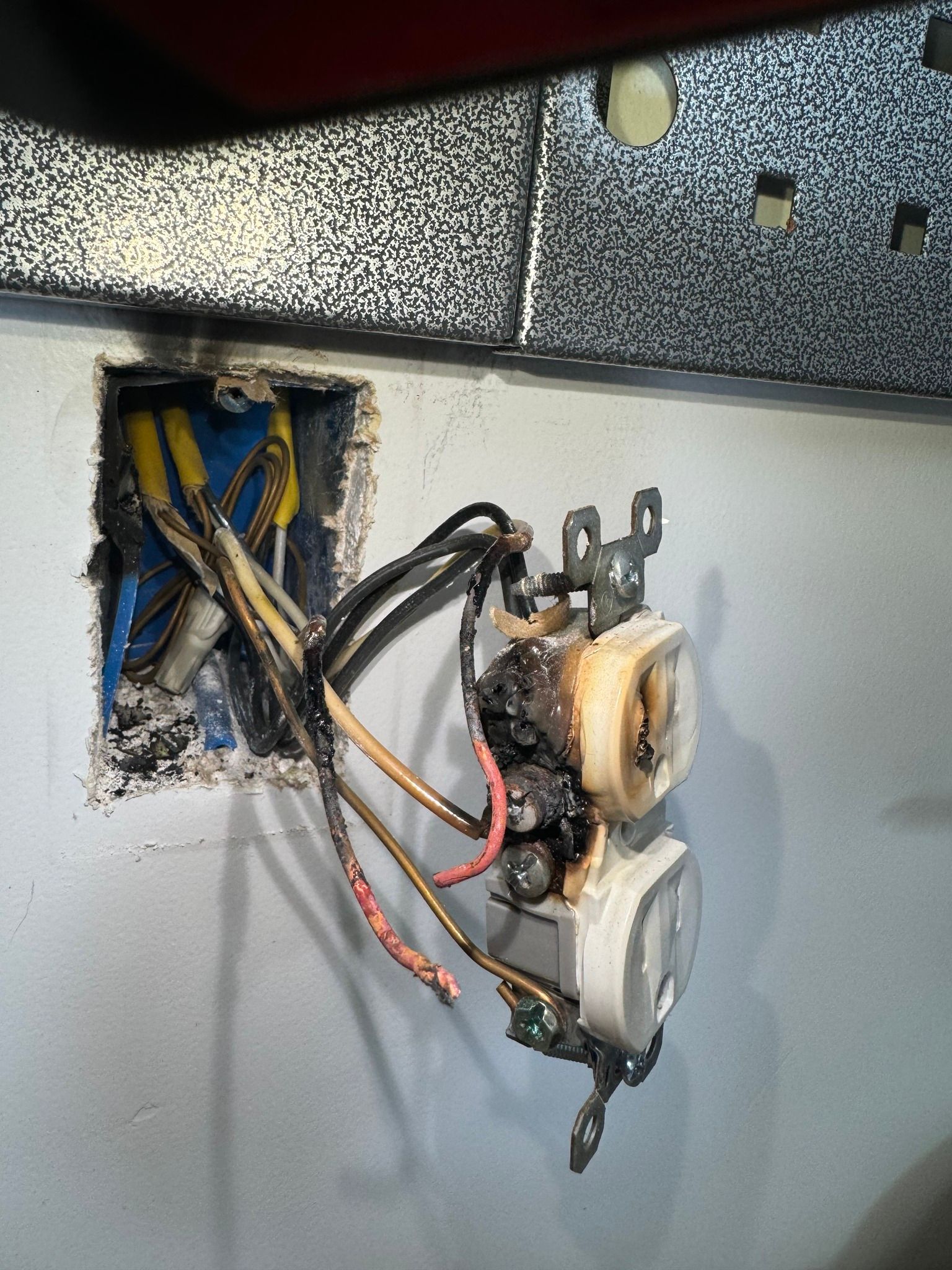 A severely damaged electrical outlet with melted, charred casing and heat-damaged wiring hanging out of a wall box.